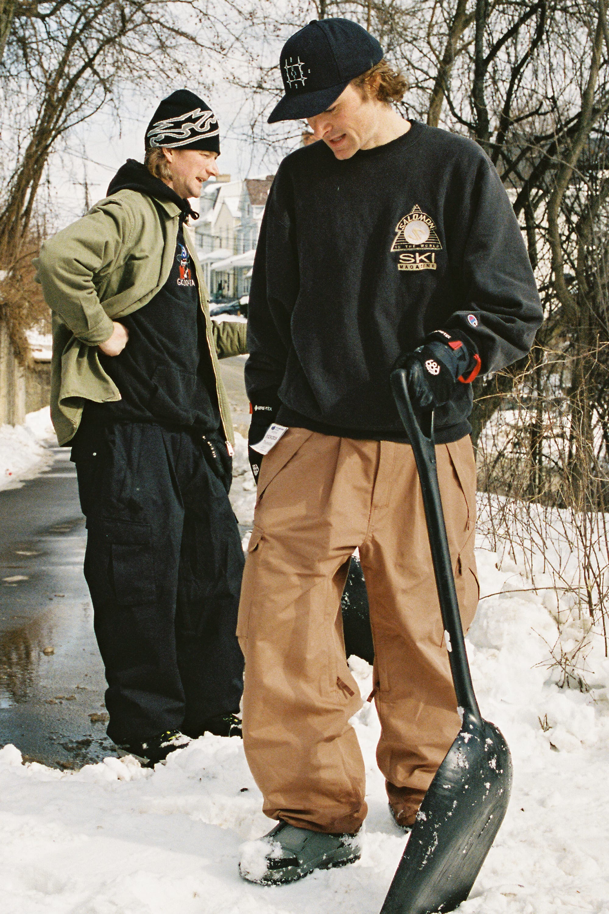 Two people stand outside in winter. One, holding a snow shovel, wears the 686 Men’s Essox™ Cargo Pant and a black sweatshirt. The other, in black clothes and a green jacket, stands nearby. Snow is on the ground; bare trees and houses are in the background.
