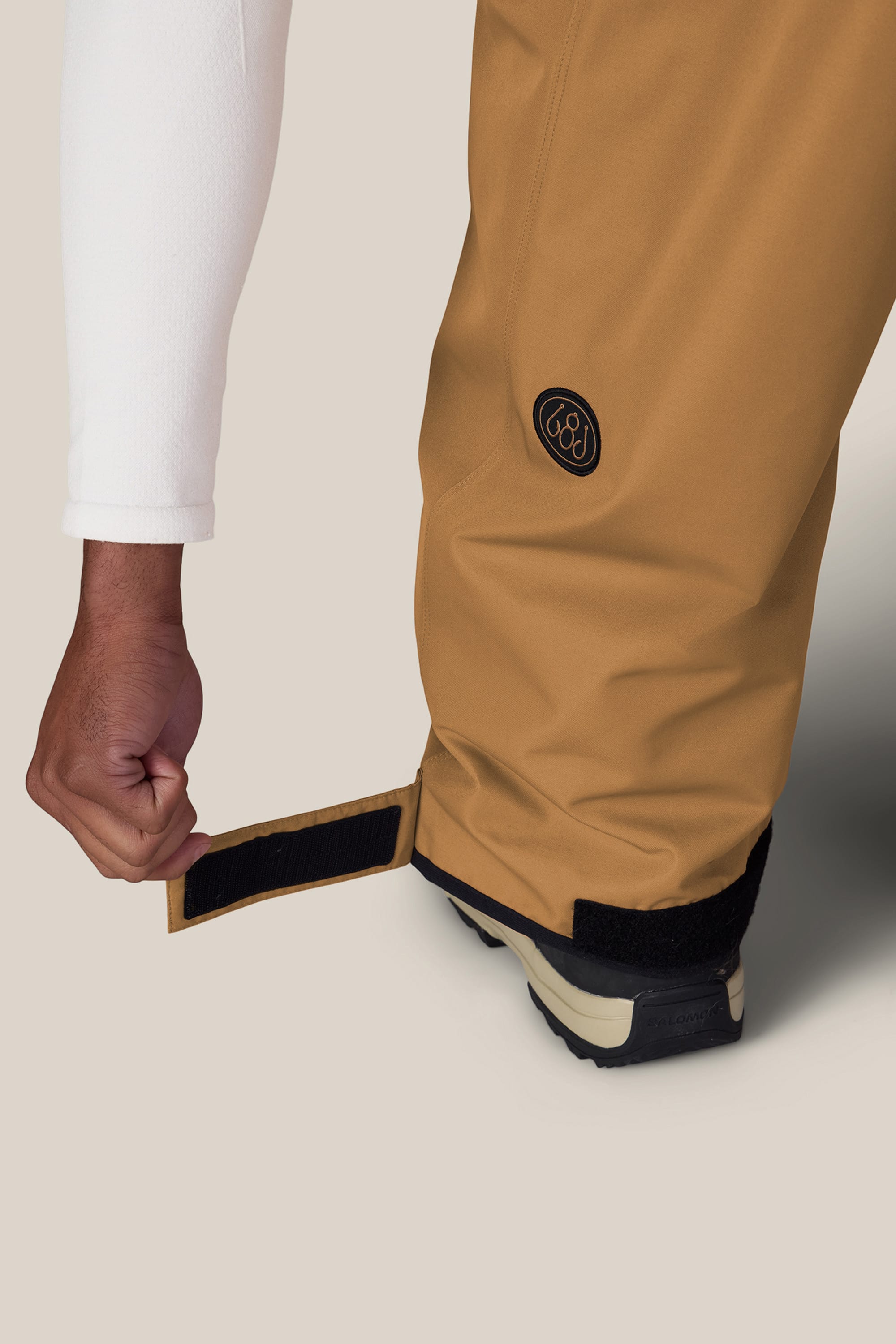A person adjusts the Velcro strap on the cuff of 686 Men’s Essox™ Cargo Pant in tan, wearing a white long-sleeve shirt and black shoes, against a plain background.