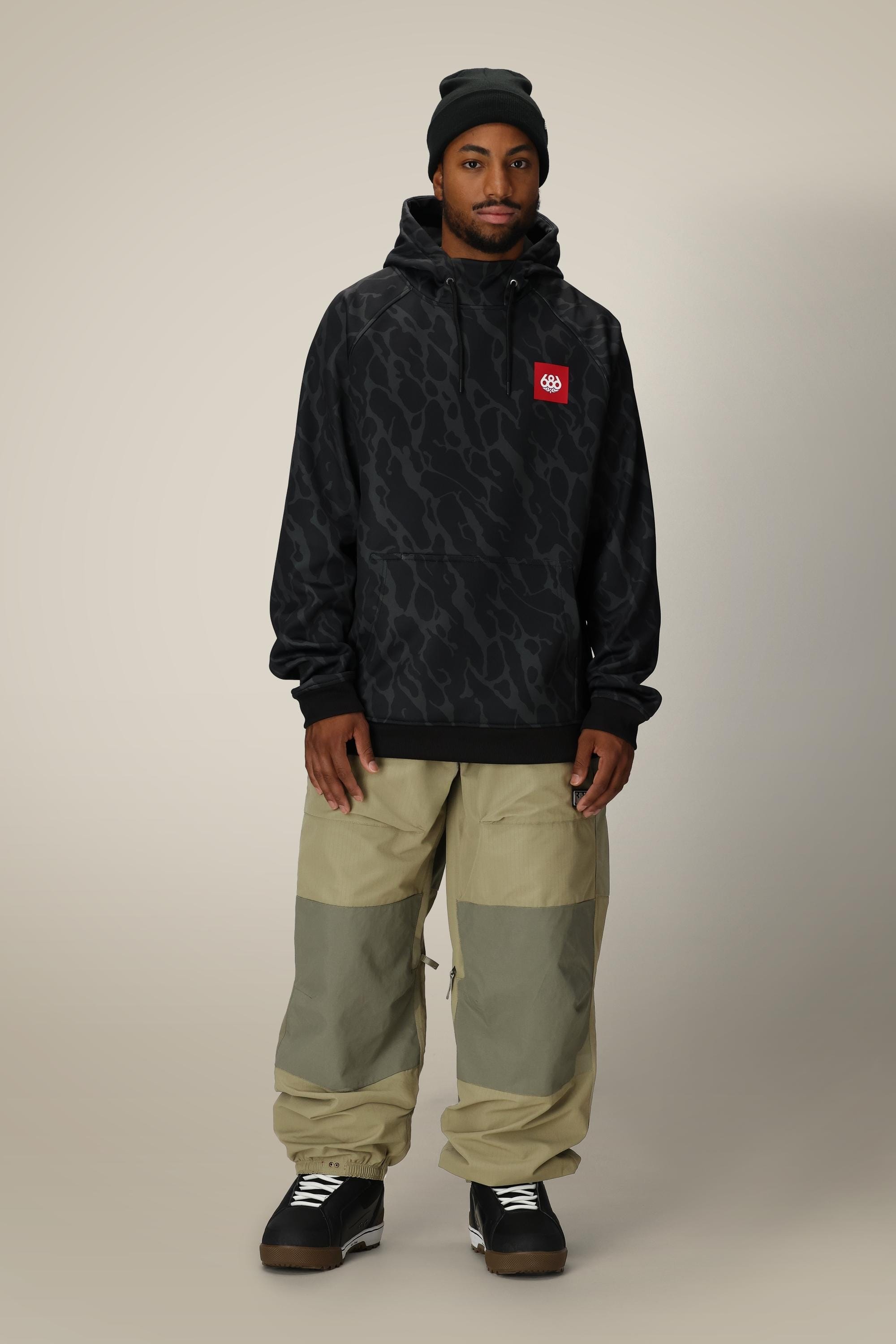 A man wearing a black beanie, 686 Mens Bonded Fleece Pullover Hoody by 686 with a red logo, light brown and olive cargo pants, and black sneakers stands against a plain beige background.