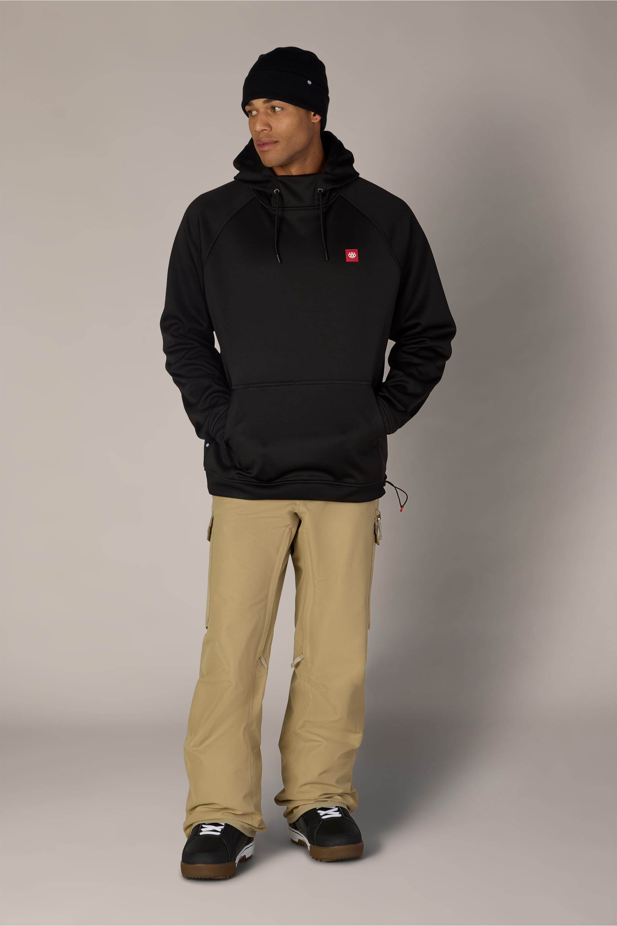 A man wearing the 686 Men’s Bonded Fleece Pullover Hoody by 686, paired with a black beanie, tan pants, and black shoes, stands with his hands in his hoodie pockets, looking to the side against a plain light background.