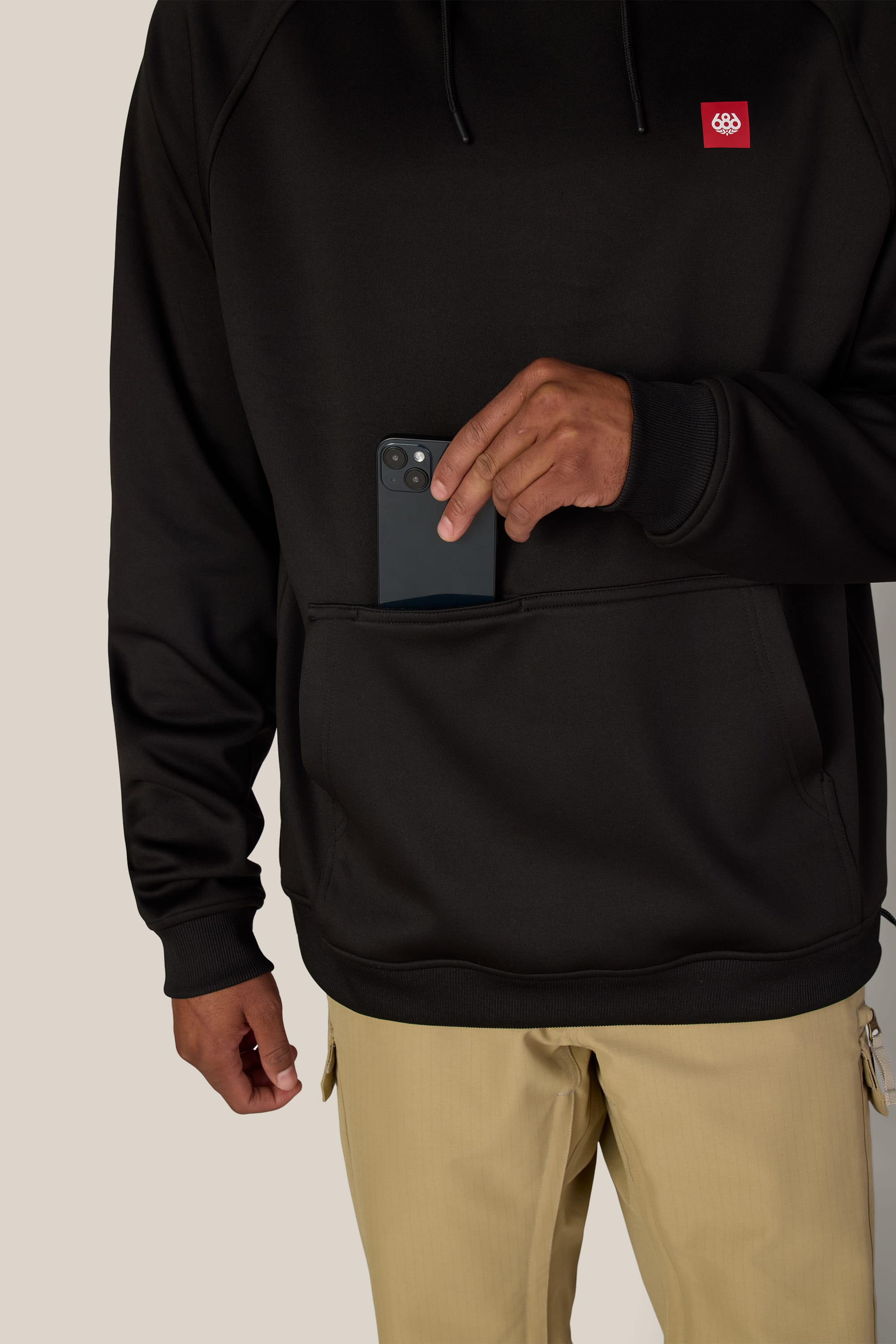 Wearing the 686 Men’s Bonded Fleece Pullover Hoody, a person in beige pants slides a black smartphone into a front pocket.