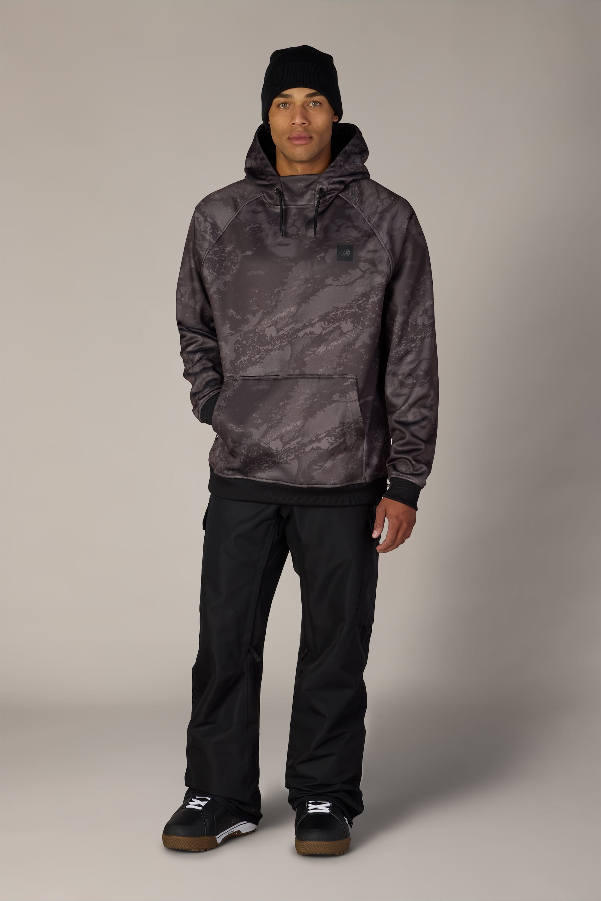 A person stands facing forward against a plain light gray background, wearing a black beanie, black pants, black sneakers, and the 686 Men’s Bonded Fleece Pullover Hoody by 686—a dark patterned hoodie—with their hands in their pockets.