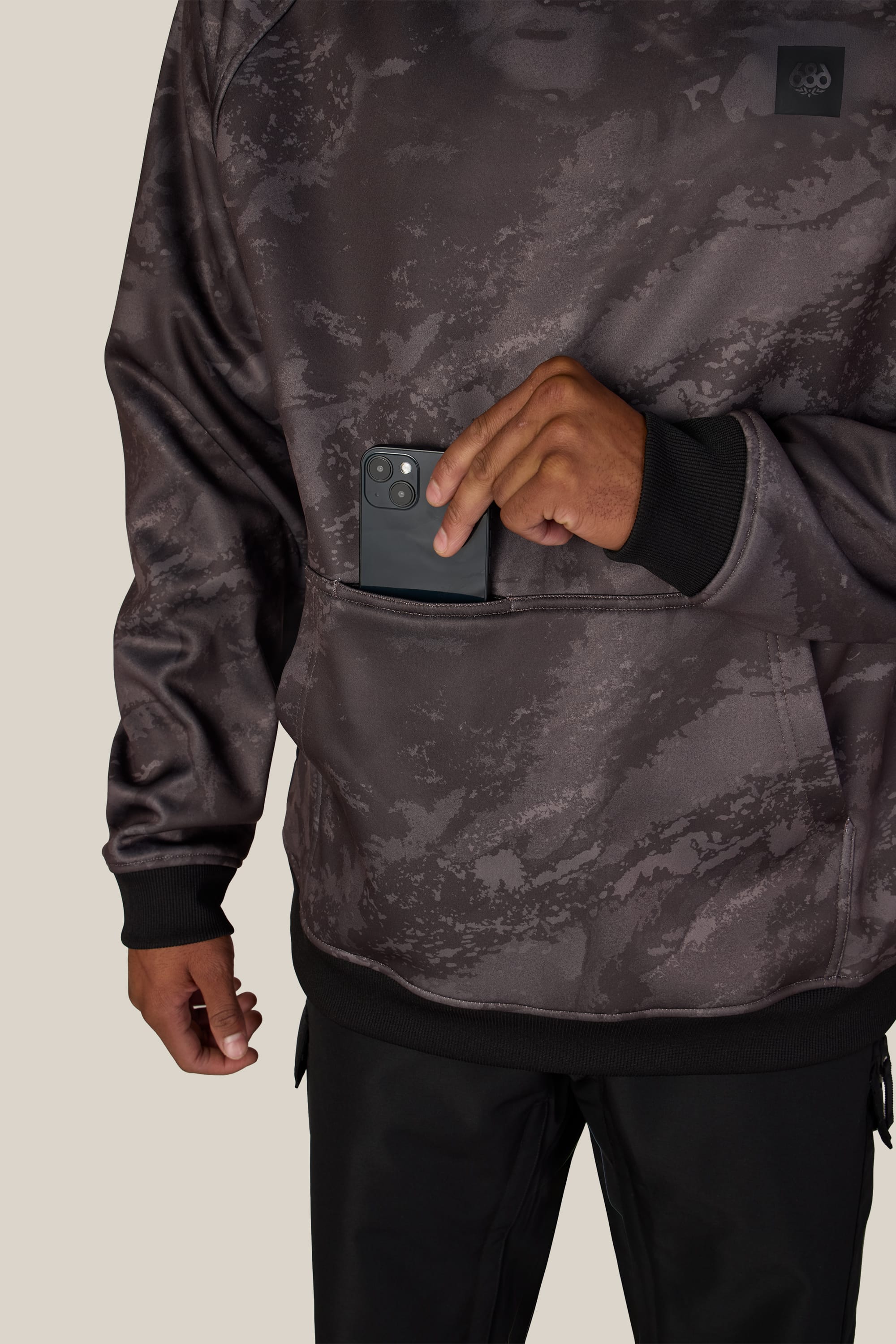 A person wearing a 686 Men’s Bonded Fleece Pullover Hoody slips a black smartphone into the front pocket. Only the lower half of their torso and one hand are visible.