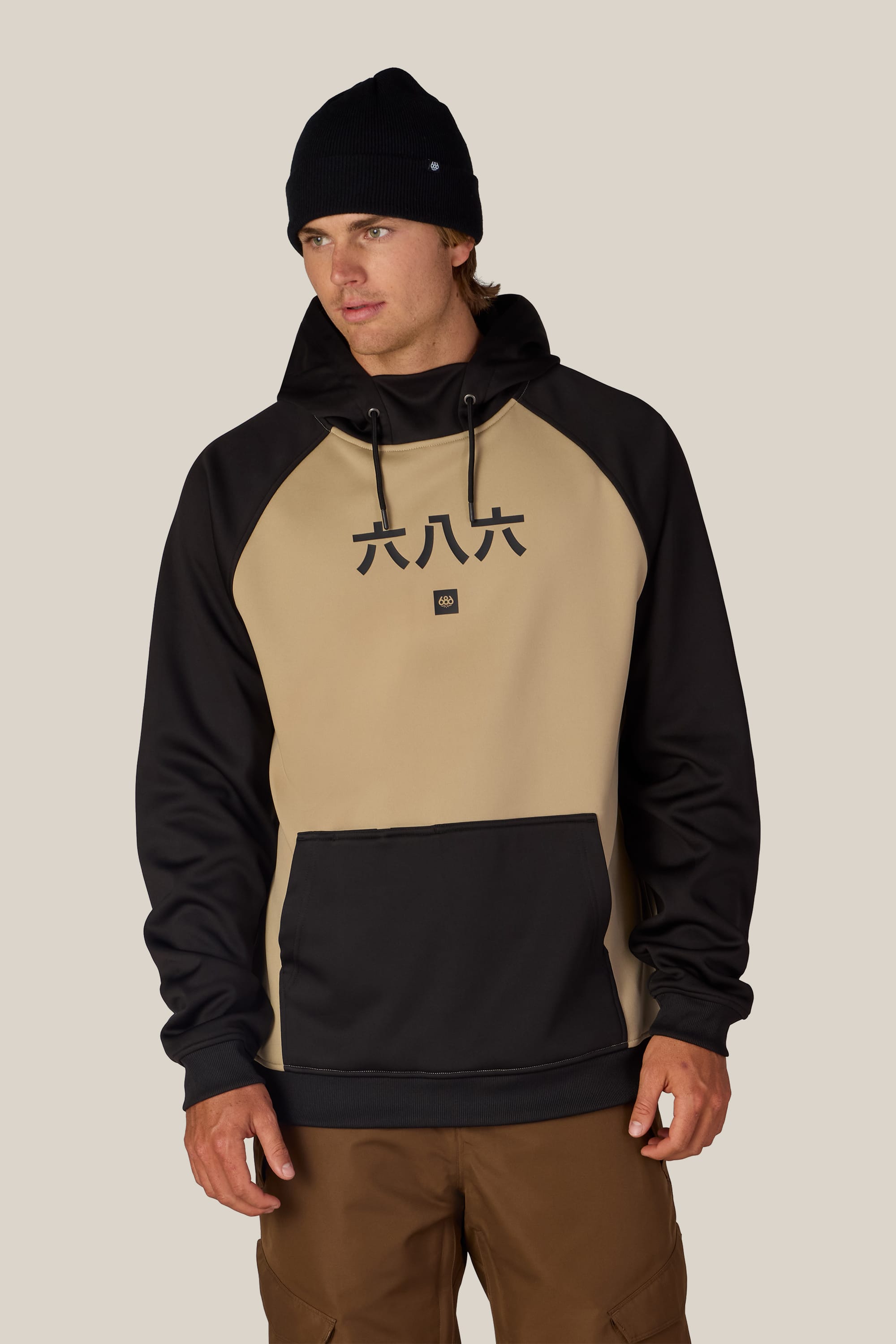 A person wearing the 686 Men’s Bonded Fleece Pullover Hoody in tan and black with Chinese characters on the front, brown pants, and a black beanie stands against a plain beige background.
