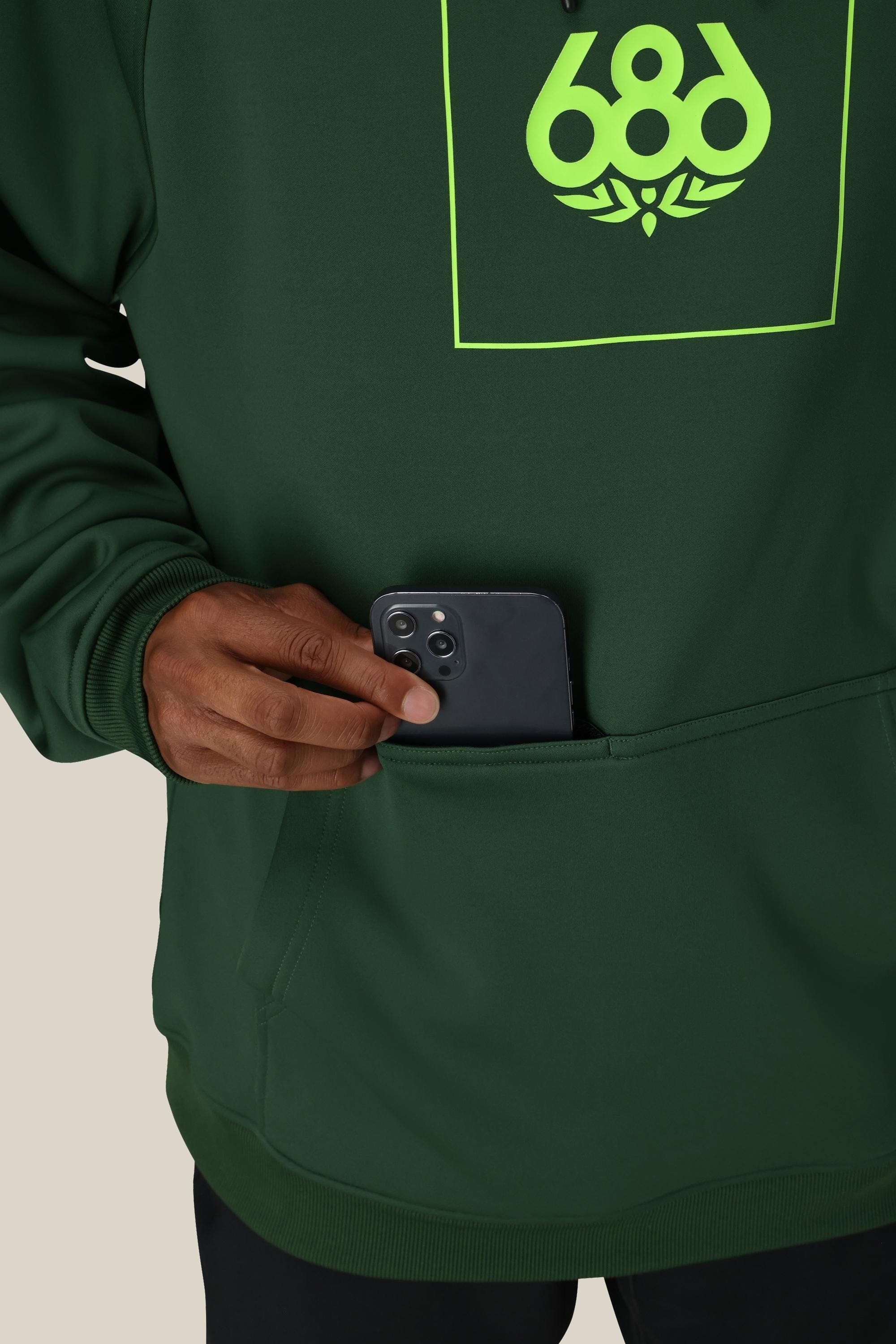 A person wearing the 686 Mens Bonded Fleece Pullover Hoody—dark green with a neon green 686 logo and cozy fleece lining—places a black smartphone into its front pocket.