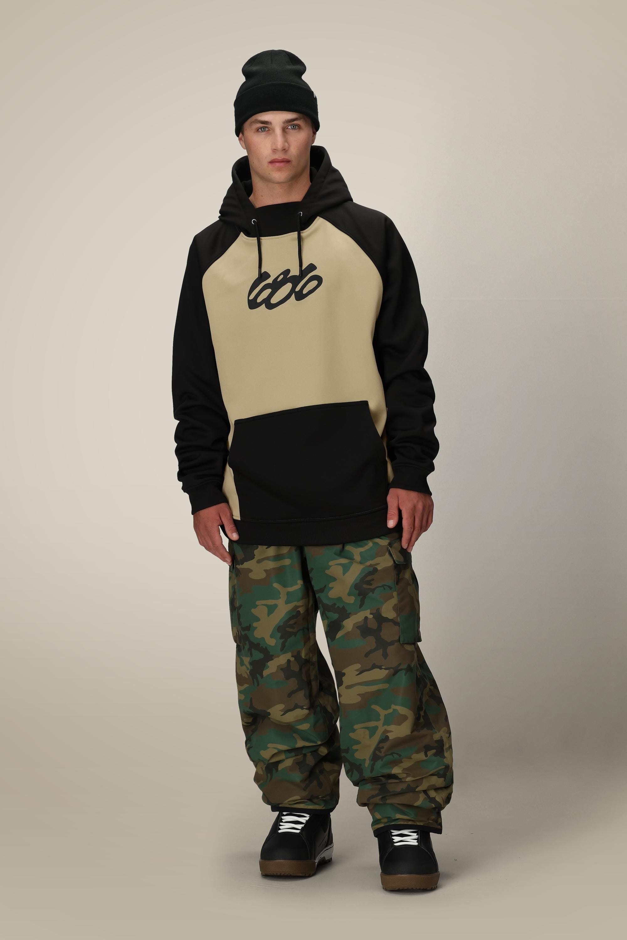 A person wears a black beanie, the 686 Mens Bonded Fleece Pullover Hoody in beige and black with 666 on the front, loose camo pants, and black shoes, standing against a plain light beige background.