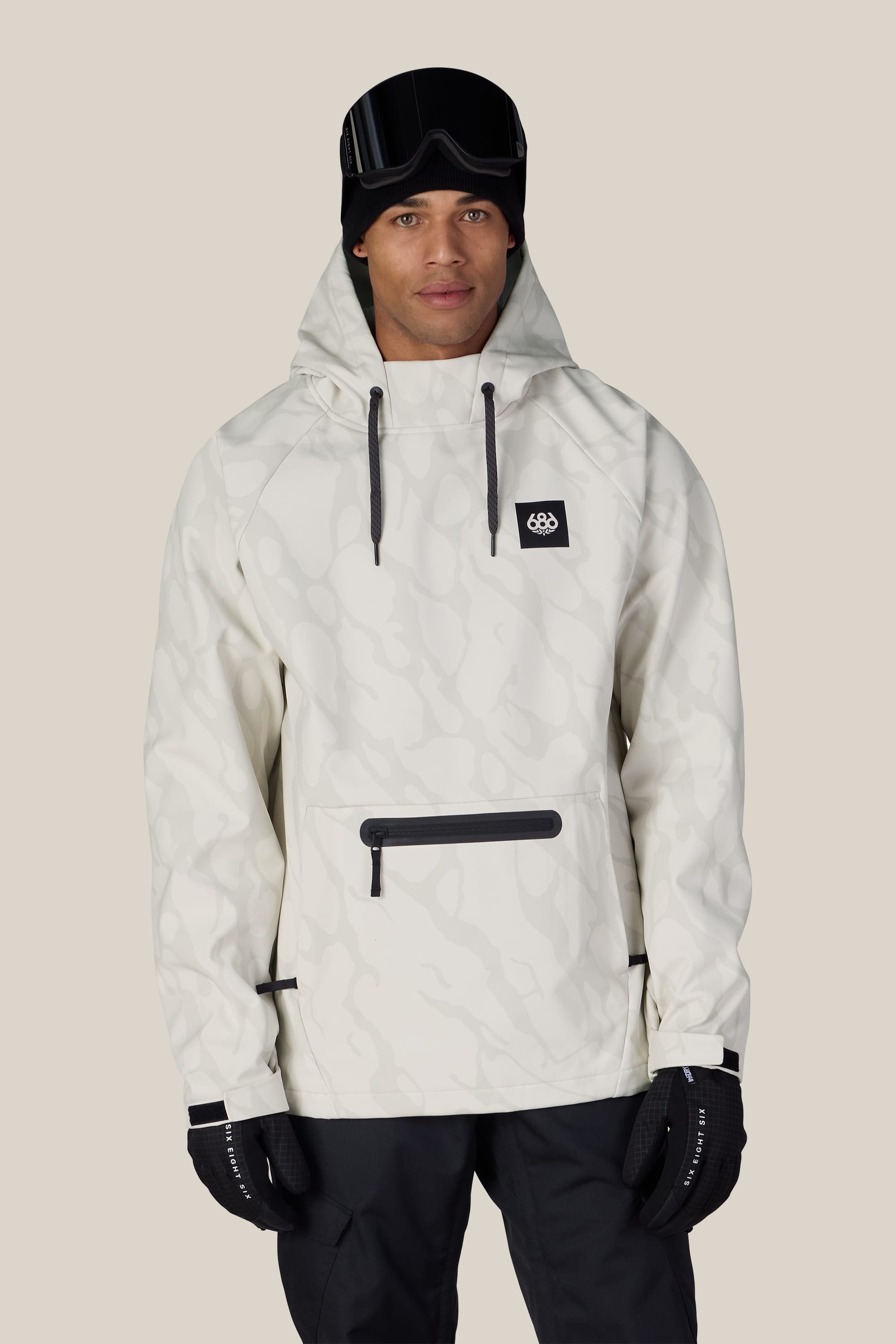 A man stands facing forward against a plain beige background, wearing the 686 Men’s Waterproof Hyperchromic Pullover Hoody with black ski goggles, gloves, and pants.