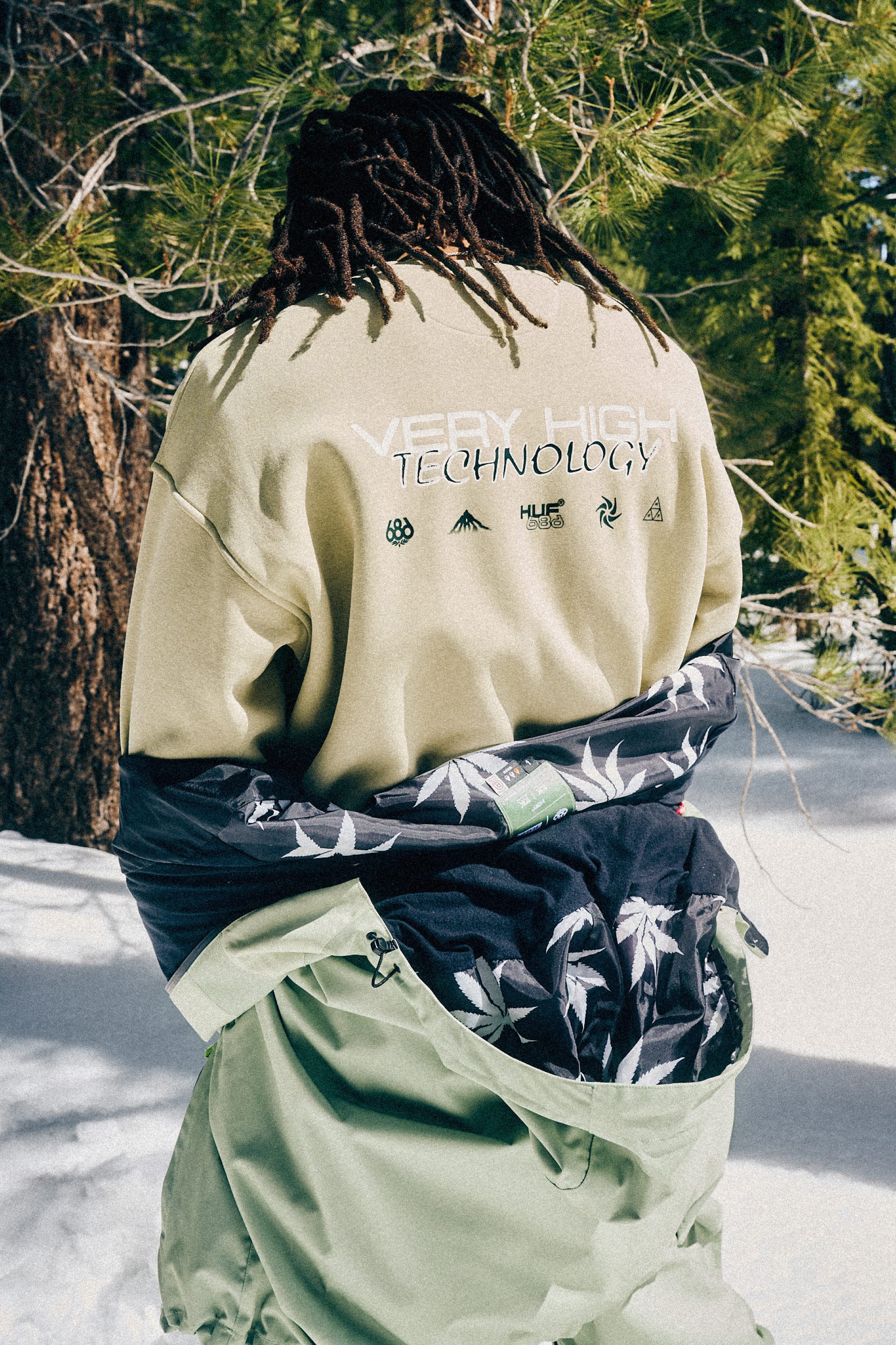 A person with dreadlocks stands in snow near trees, wearing a light green men’s cotton blend jacket and pants, with the jacket partly off to reveal the 686 HUF Premium Heavyweight Crew Sweatshirt by 686.