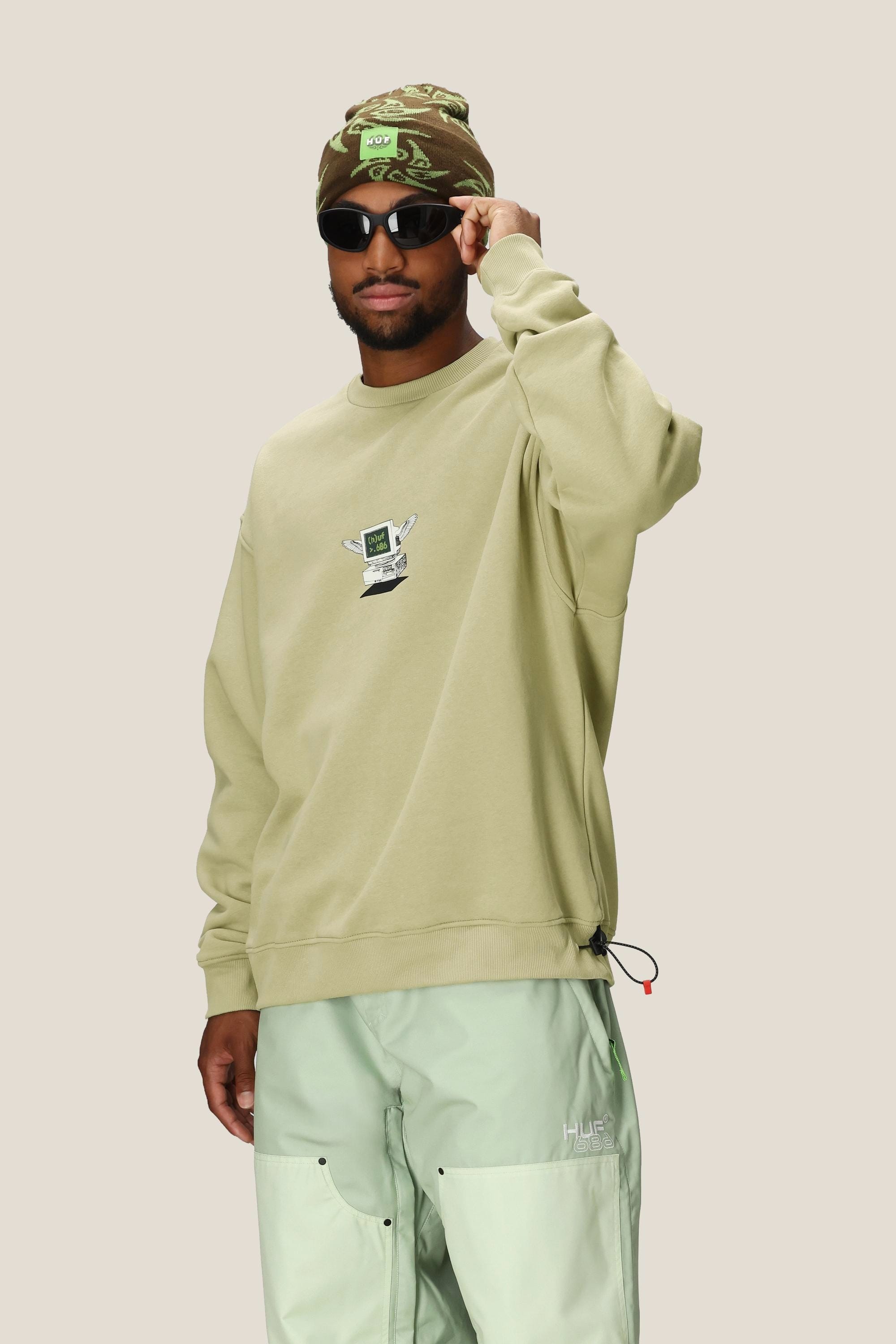 A man wearing sunglasses, a camouflage beanie, and the 686 HUF Premium Heavyweight Crew Sweatshirt by 686 in light green, paired with pale mint green pants, stands against a plain beige background.