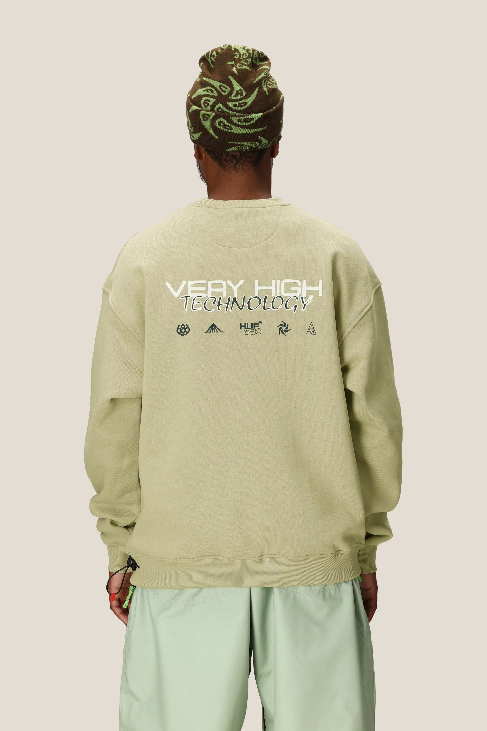 A person stands facing away, wearing a light green 686 HUF Premium Heavyweight Crew Sweatshirt with graphics on the back, mint green shorts, and a brown-green patterned beanie. The background is solid light beige.