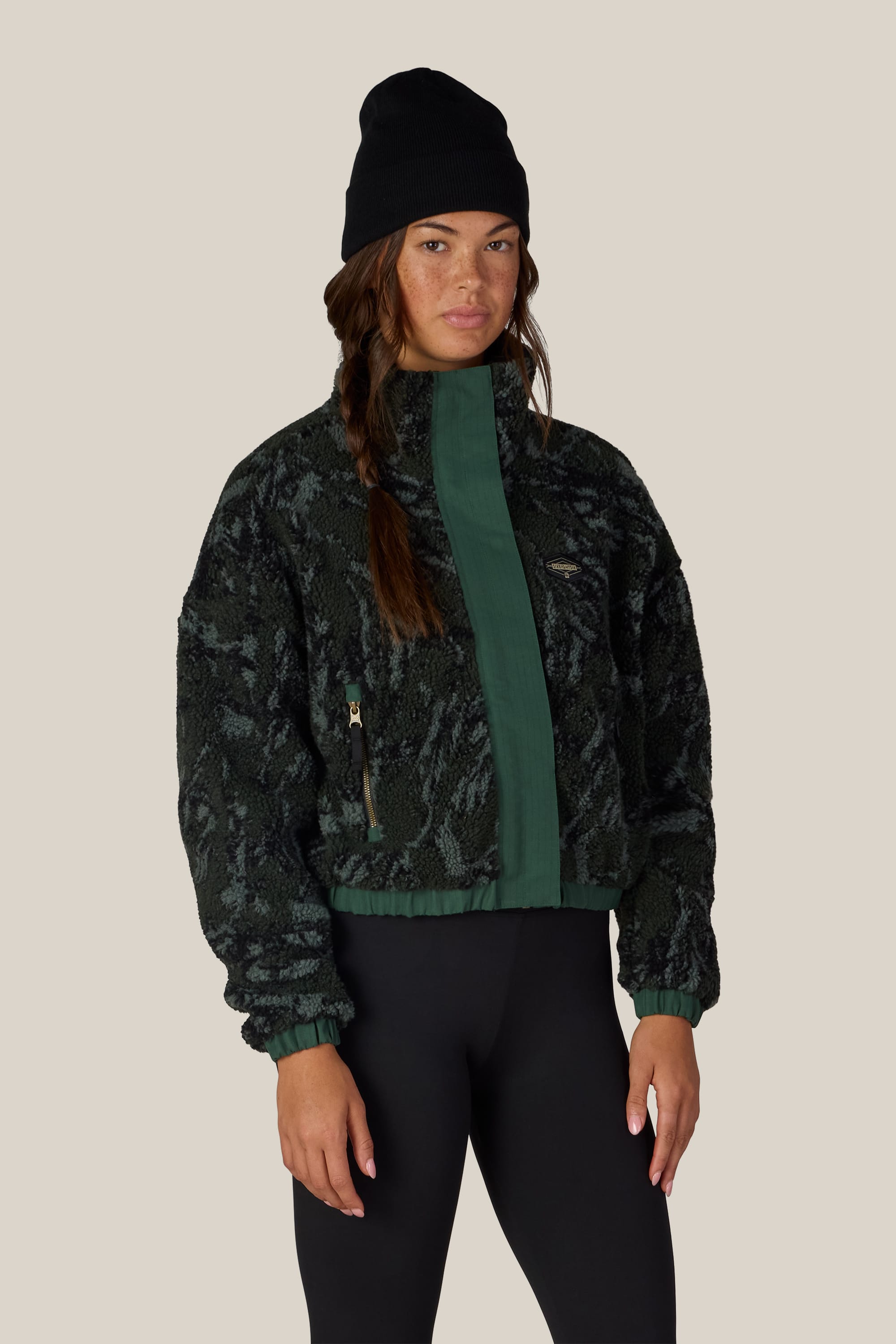 A person wearing a black beanie, the 686 Women’s Outpost Sherpa Jacket by 686 in a green and black textured design, and black leggings stands against a plain beige background.