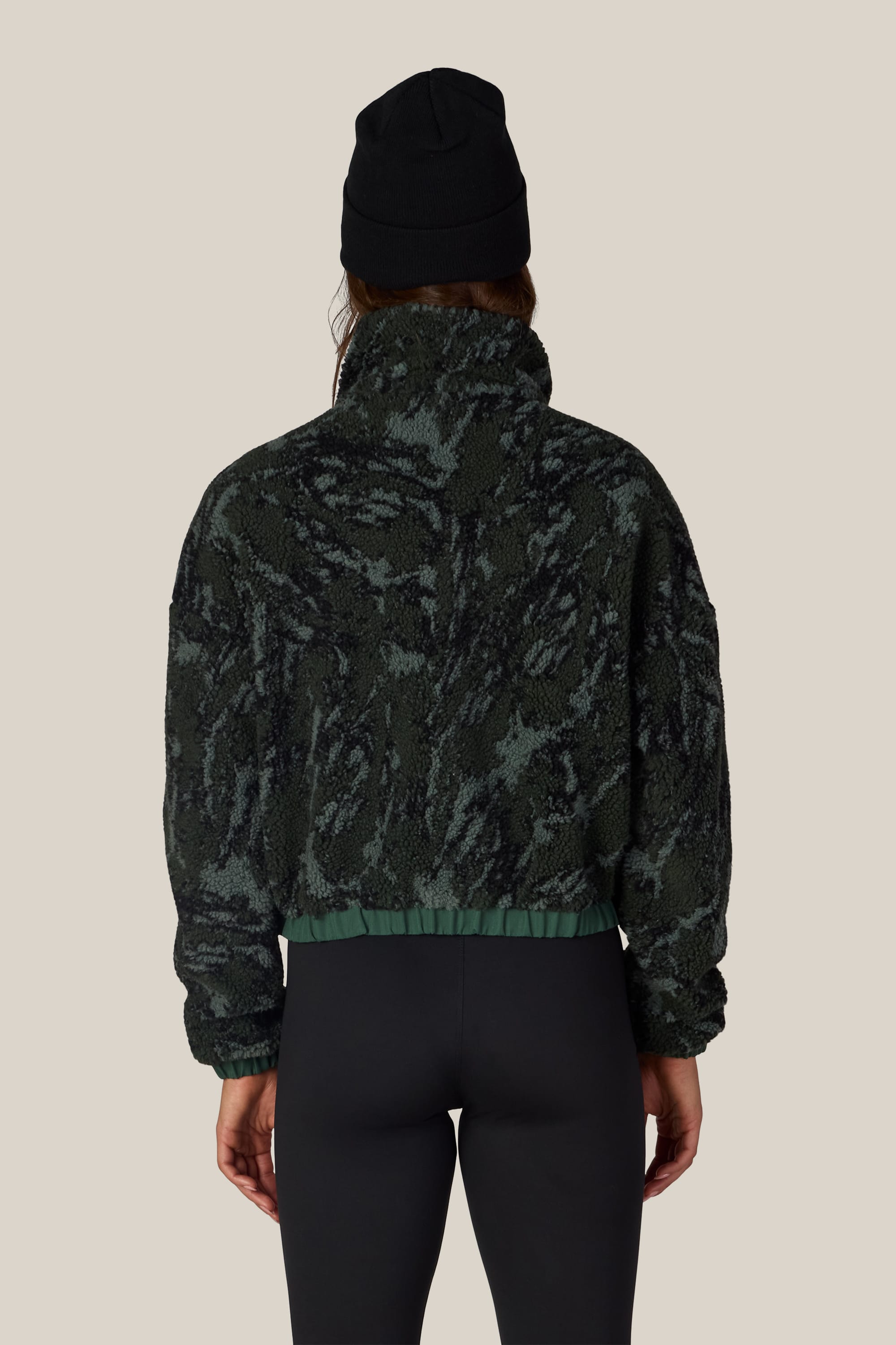 A person is seen from behind against a light beige background, wearing the 686 Women’s Outpost Sherpa Jacket in a dark green pattern by 686, paired with black leggings and a black beanie.