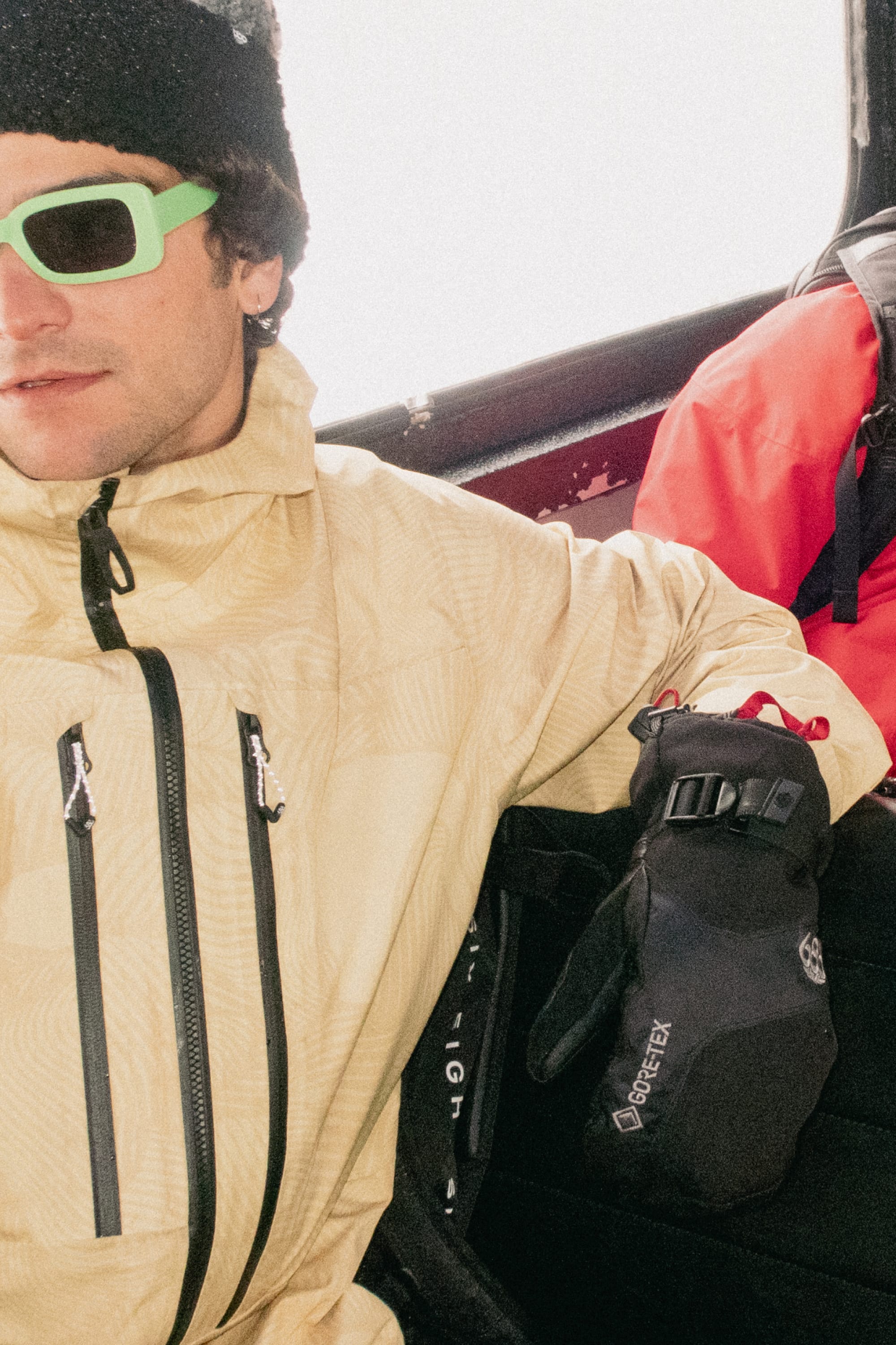 A person in a yellow jacket, 686 Men's GORE-TEX Leather Linear™ Mitt by 686, green sunglasses, and a black beanie sits next to a red backpack, likely on a ski lift or tram, with bright light in the background.