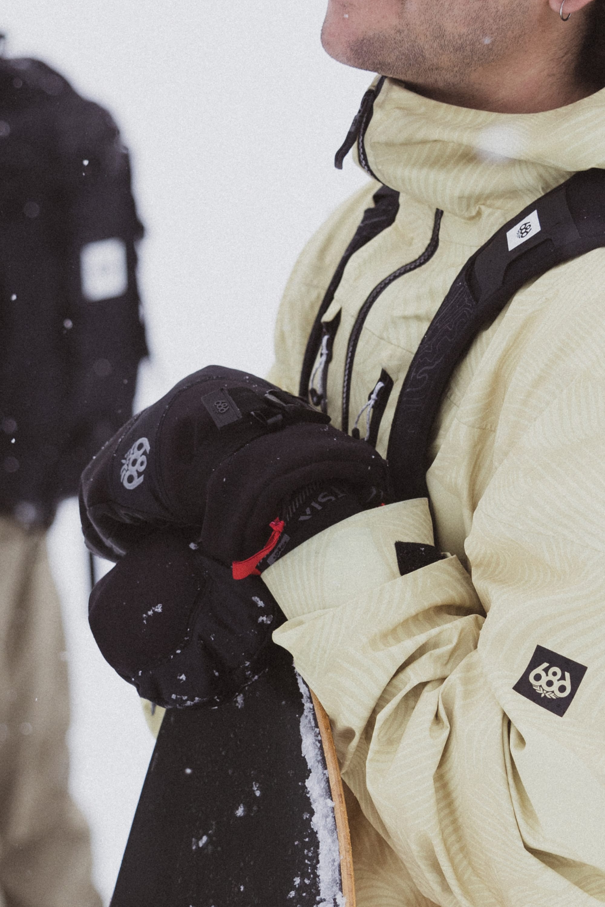 A person in a light jacket wears 686 Men's GORE-TEX Leather Linear™ Mitts and holds a snowboard. Their face is partially out of frame with visible snowflakes, while someone in dark clothing is blurred in the background.