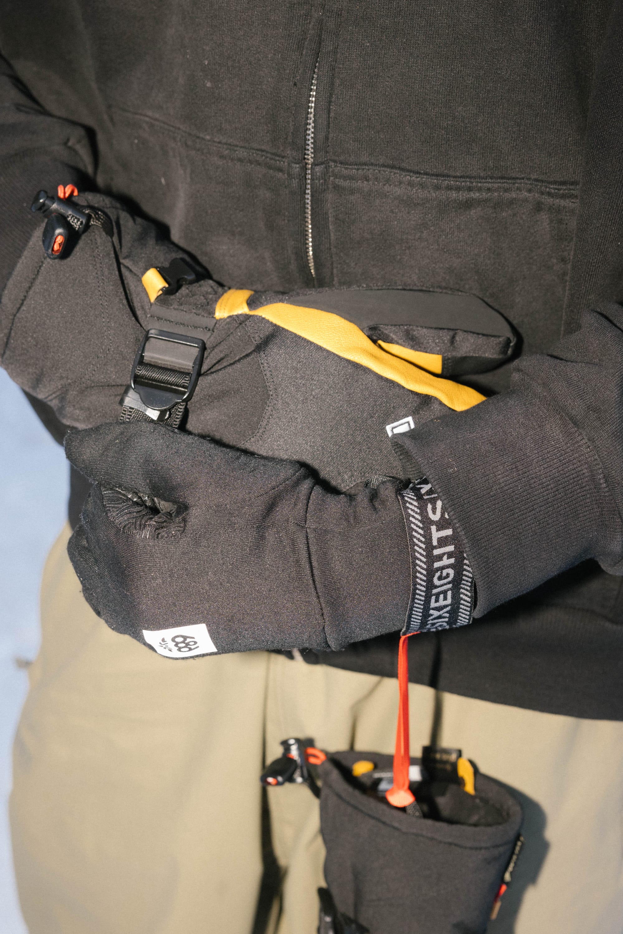 Close-up of a person wearing 686 Men's GORE-TEX Leather Linear™ Mitts with black leather palms and black gloves, adjusting a black and yellow strap on their wrist. They're in a snowy setting, dressed in a black jacket and beige pants. Brand: 686.