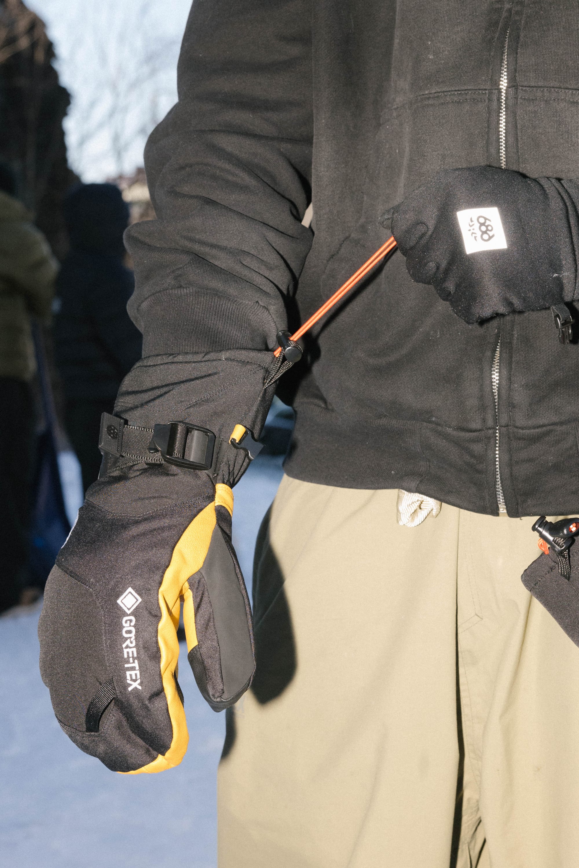 Wearing winter clothes, a person stands outdoors on snow and uses a tool to adjust their black and yellow 686 Men’s GORE-TEX® Leather Linear™ Mitt.
