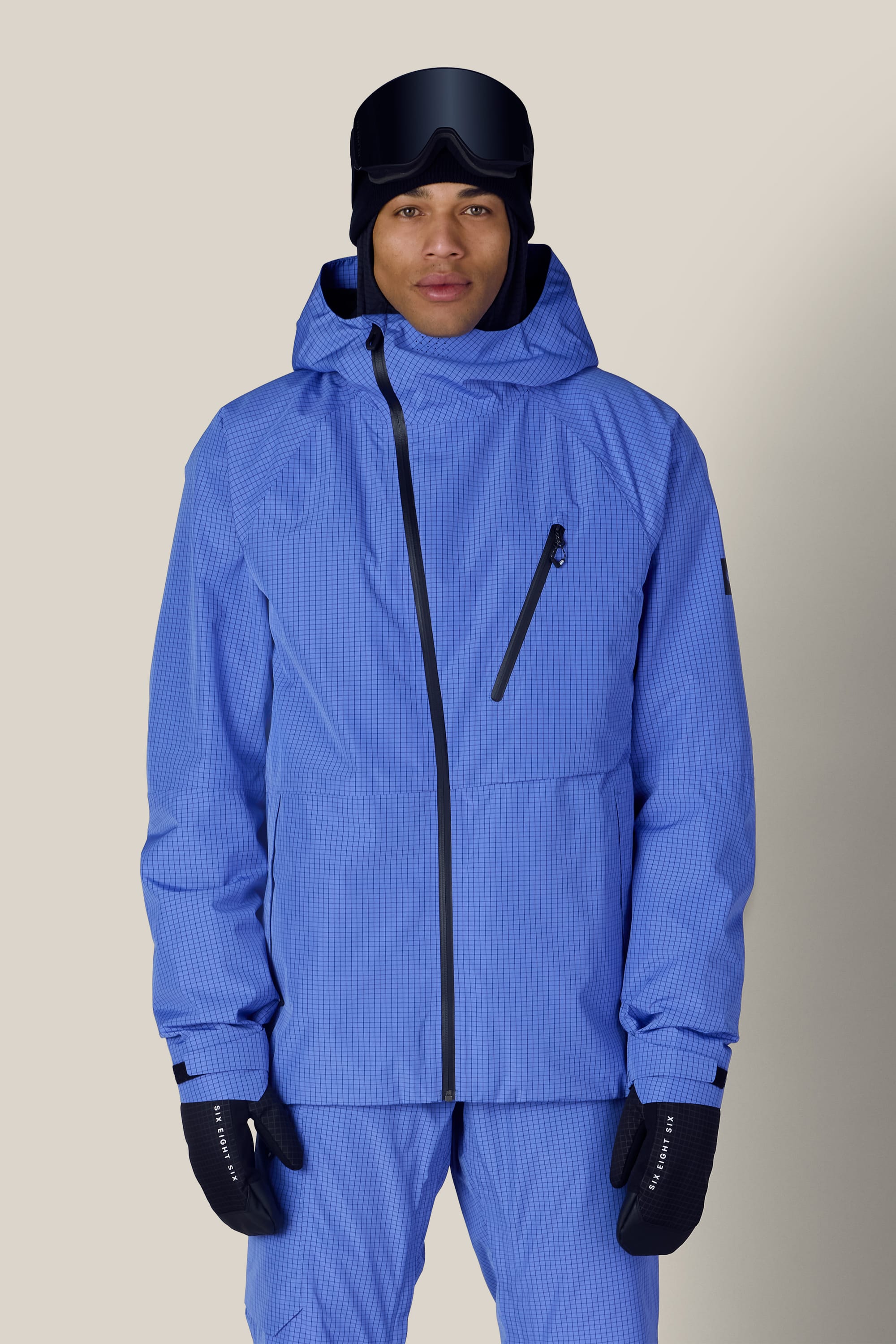 A person wearing the 686 Men’s Hydra™ Thermagraph® Jacket by 686, along with matching pants, black gloves, a black beanie, and ski goggles, stands against a light beige background.