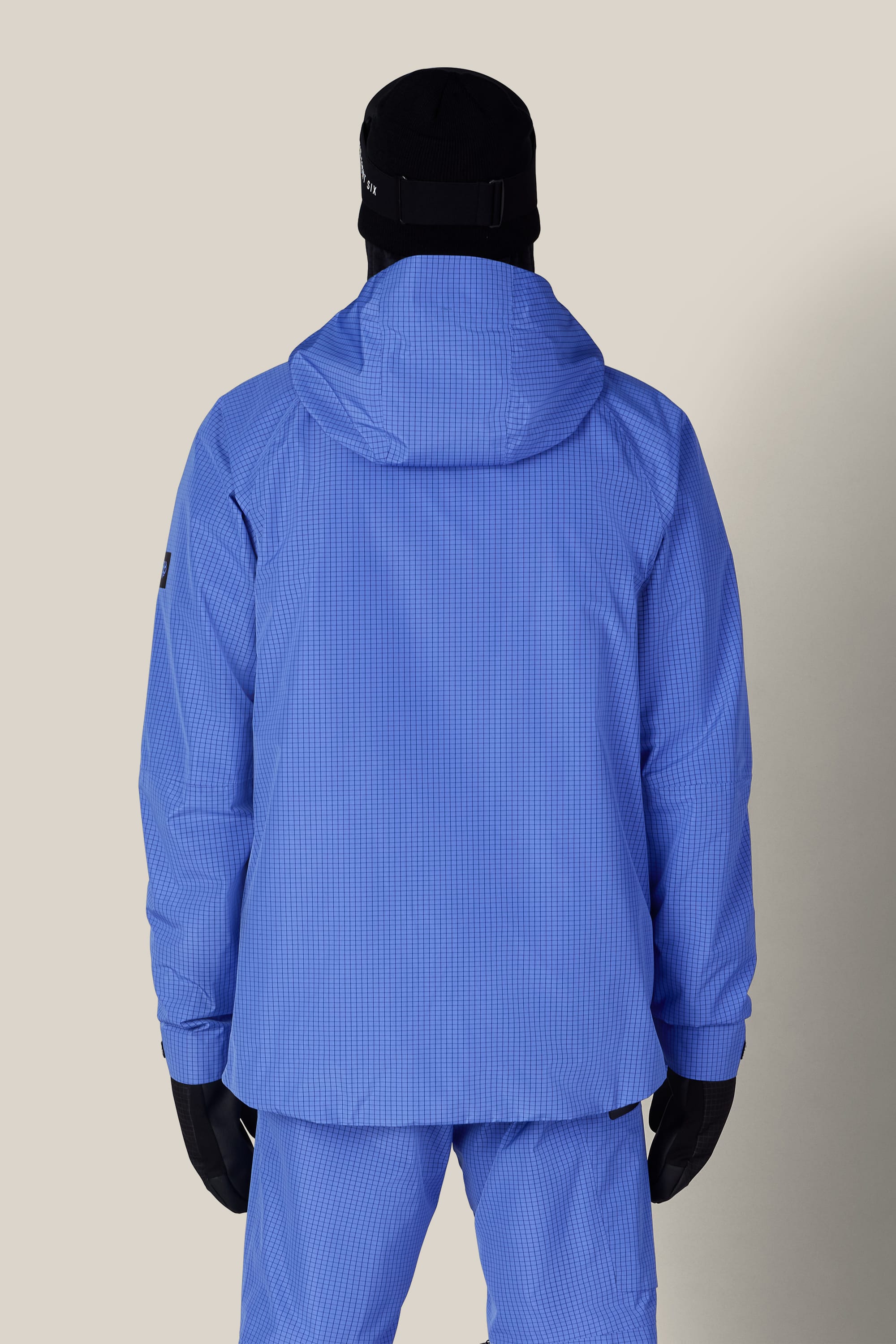 A person is seen from behind, wearing the 686 Men’s Hydra™ Thermagraph® Jacket in blue along with matching pants, black gloves, and a black hat, standing against a plain beige background.