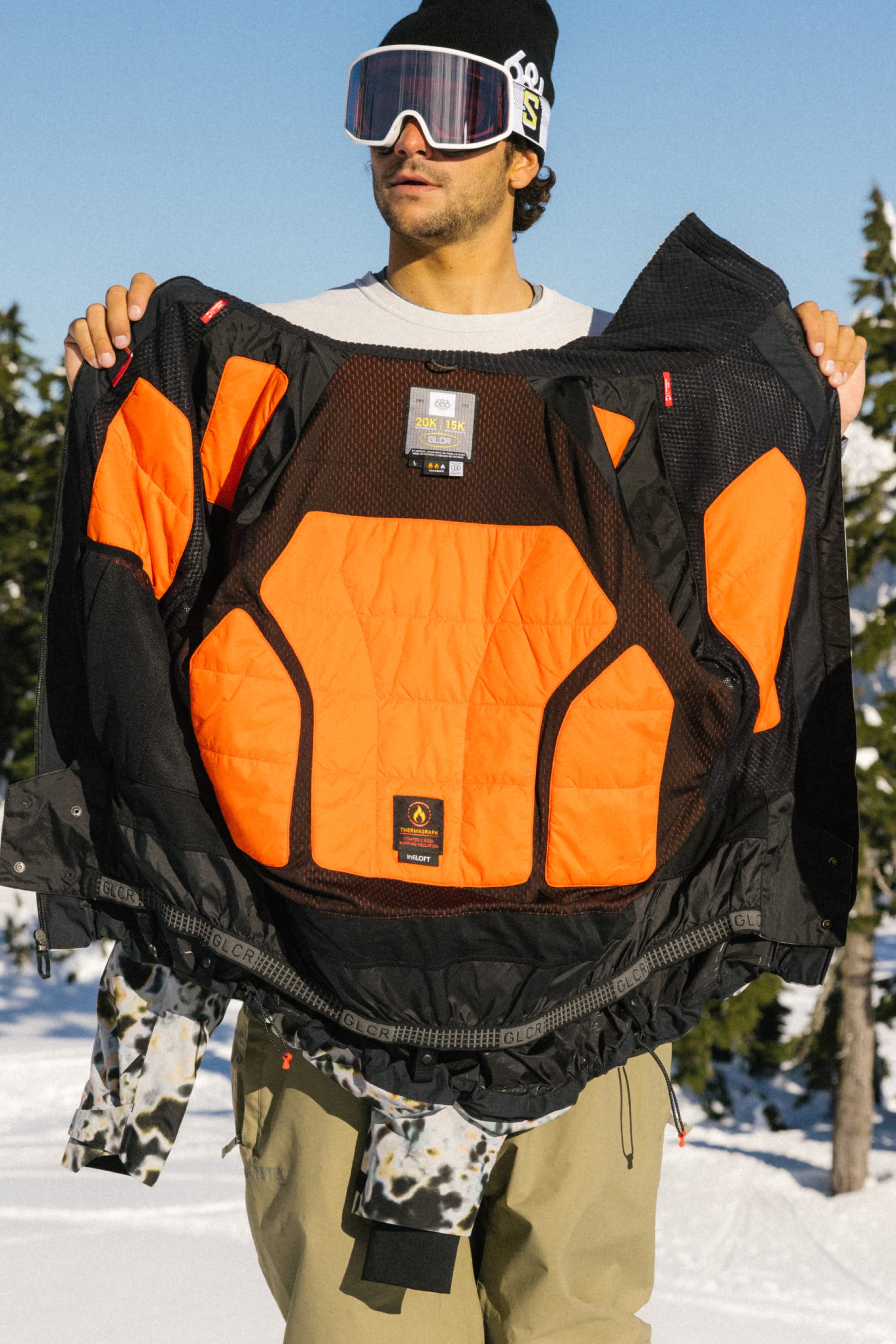 Wearing ski goggles and winter gear, a person stands in a snowy forest holding open the 686 Men's Hydra™ Thermagraph® Jacket by 686, revealing its bright orange body-mapped lining and interior labels.