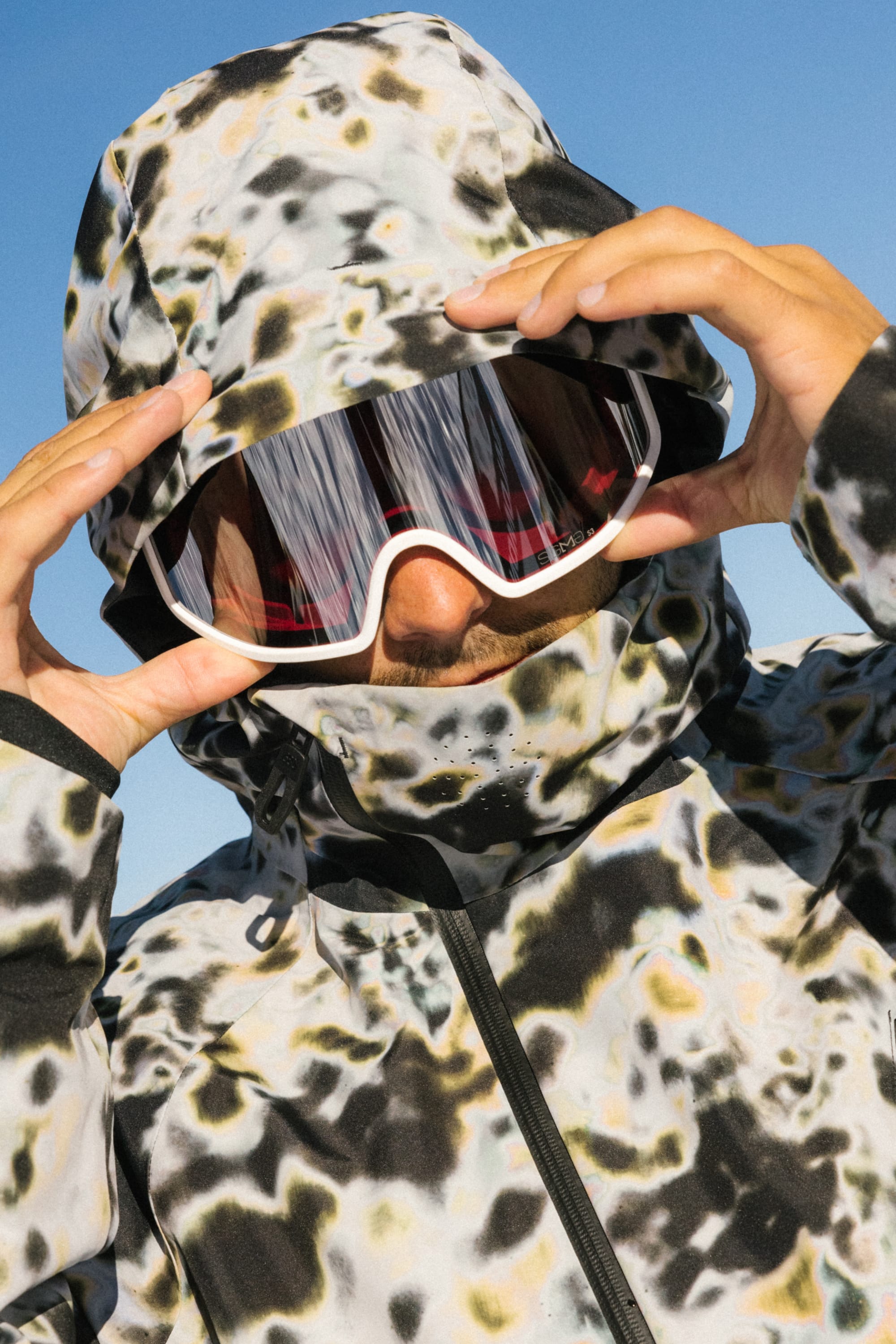 A person adjusts the hood of the 686 Men's Hydra™ Thermagraph® Jacket, a camo-patterned, do-it-all layer from 686, while wearing reflective ski goggles under a clear blue sky. Their face is mostly hidden by the hood and goggles.