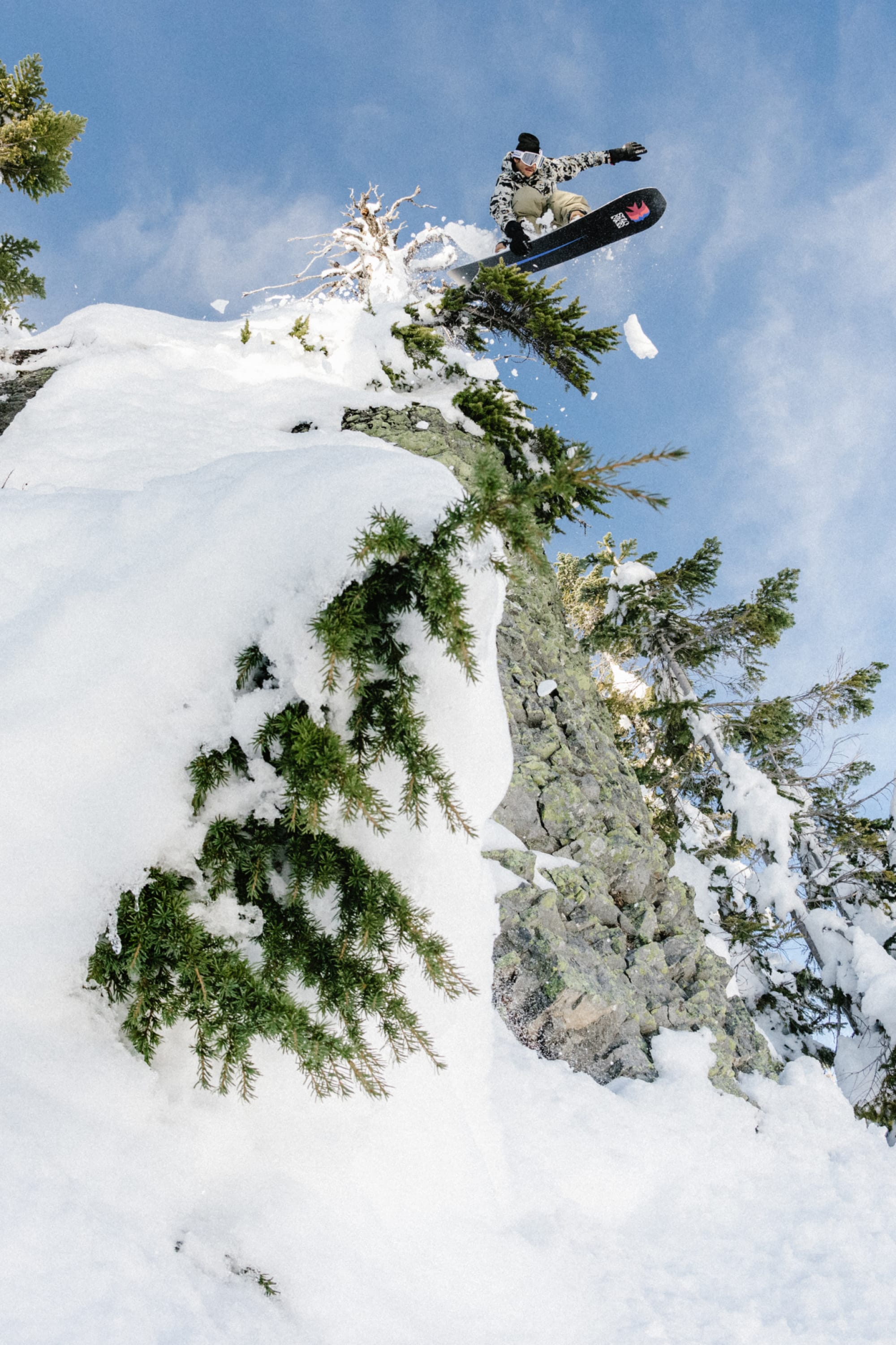 Wearing the 686 Men's Hydra™ Thermagraph® Jacket, a snowboarder soars mid-air off a snowy, tree-lined cliff under a blue sky, snow spraying behind—a perfect display of the 686 jacket’s all-mountain performance.