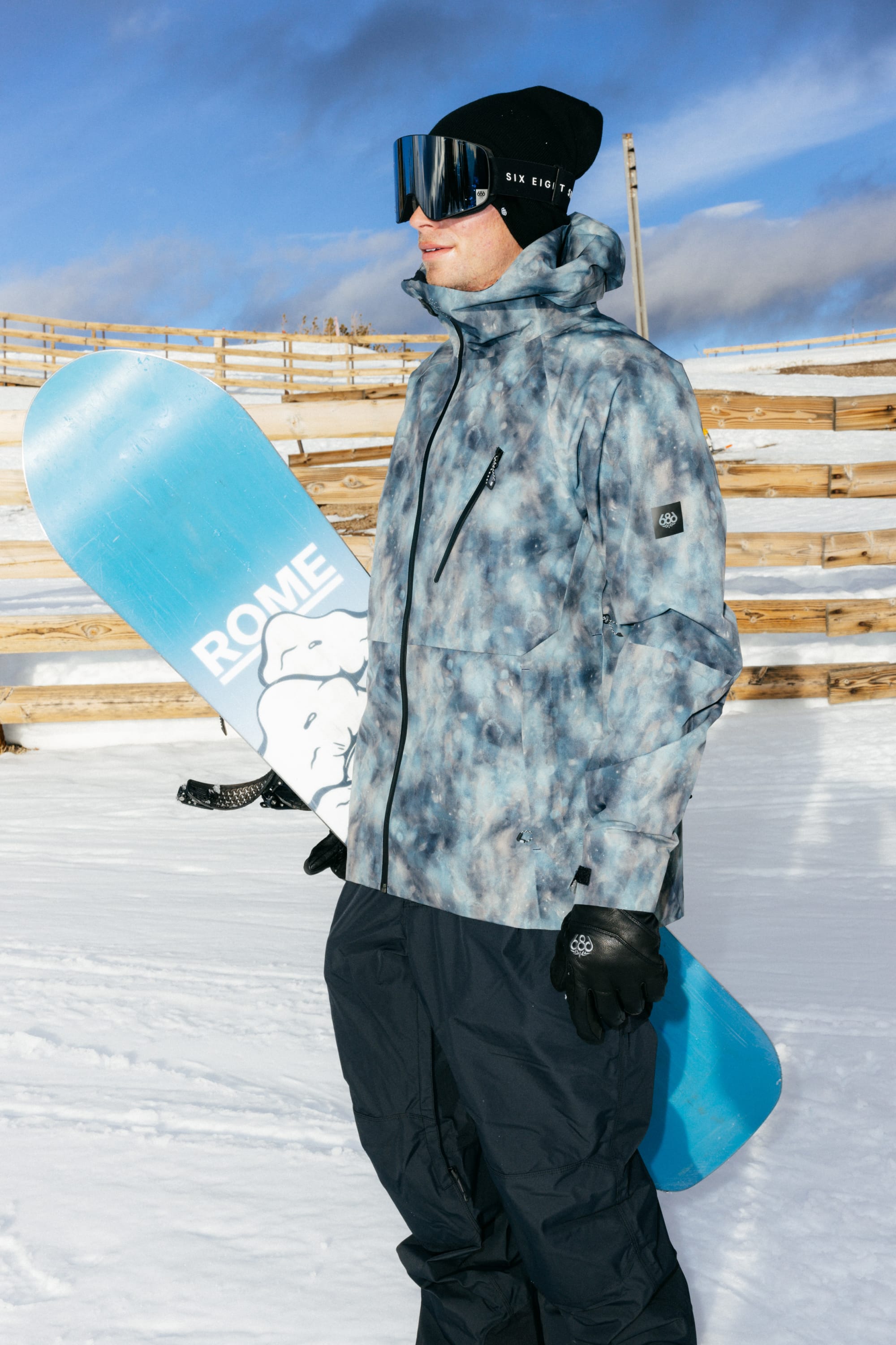 A person wearing the 686 Men’s Hydra™ Thermagraph® Jacket stands on snow with goggles, holding a blue snowboard labeled ROME, against a backdrop of wooden fences and a blue sky.