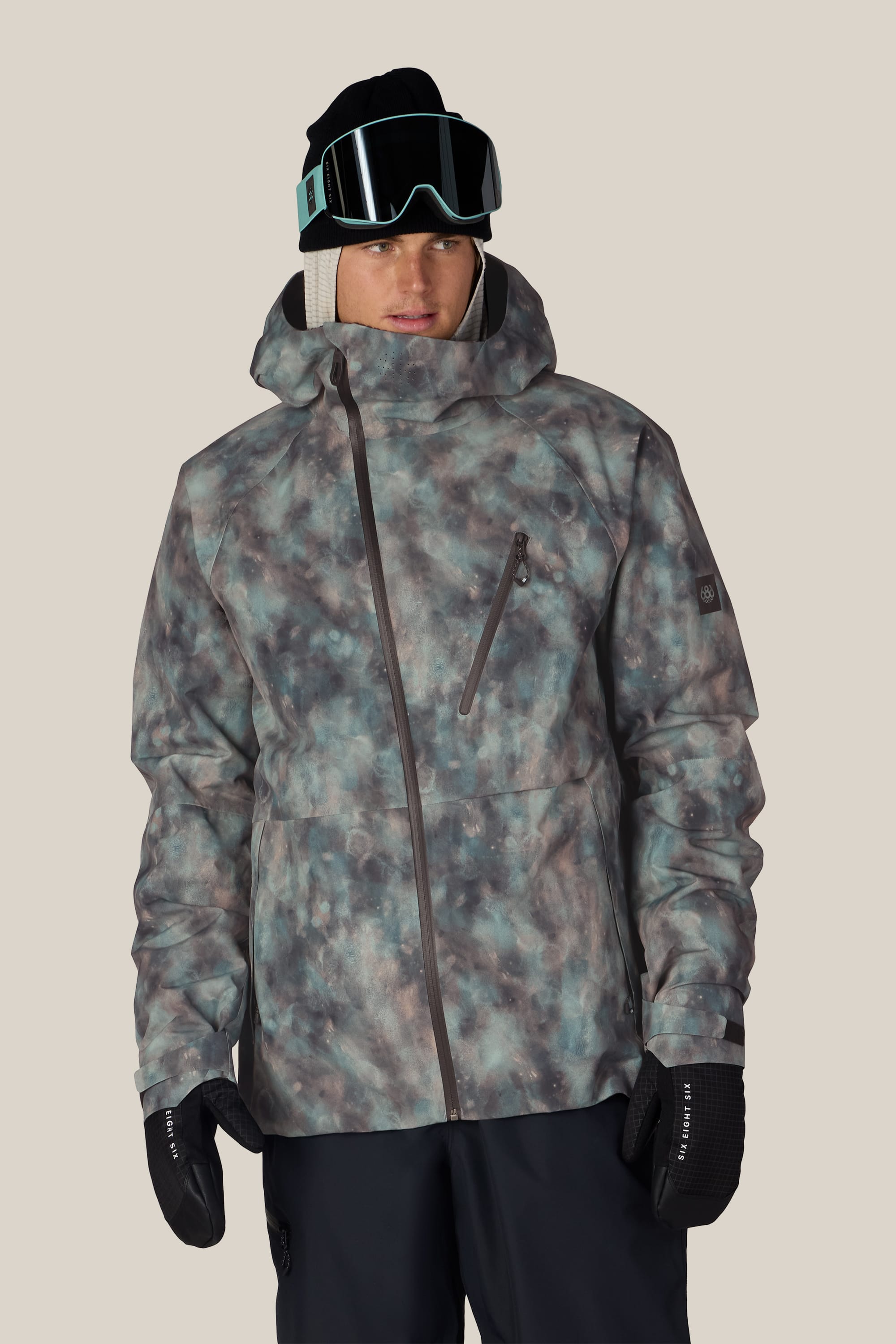 A person wearing the 686 Men’s Hydra™ Thermagraph® Jacket by 686, paired with black snow pants, black gloves, dark ski goggles, and a black beanie, stands against a plain light background.