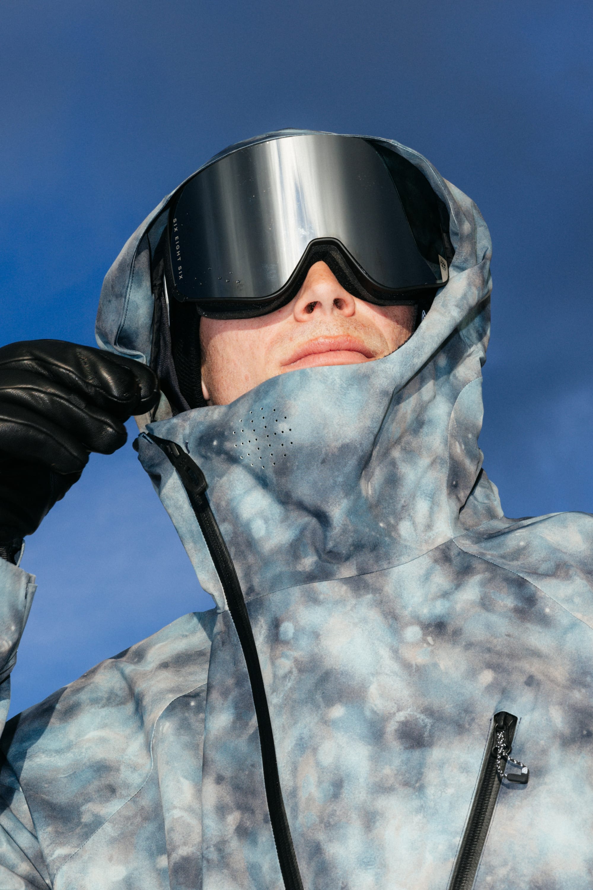 A person wearing the 686 Men’s Hydra™ Thermagraph® Jacket by 686, along with black gloves and large ski goggles, stands outdoors against a blue sky, adjusting their hood.