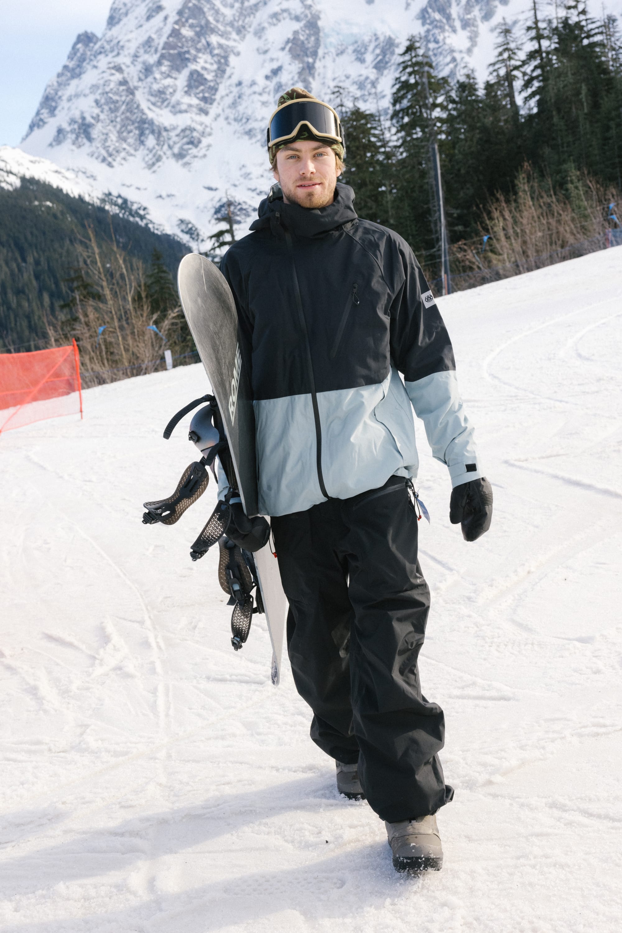 A snowboarder in a 686 Men's Hydra™ Thermagraph® Jacket, black pants, gloves, and goggles walks on a snowy slope carrying a snowboard, with pine trees and mountains in the background.