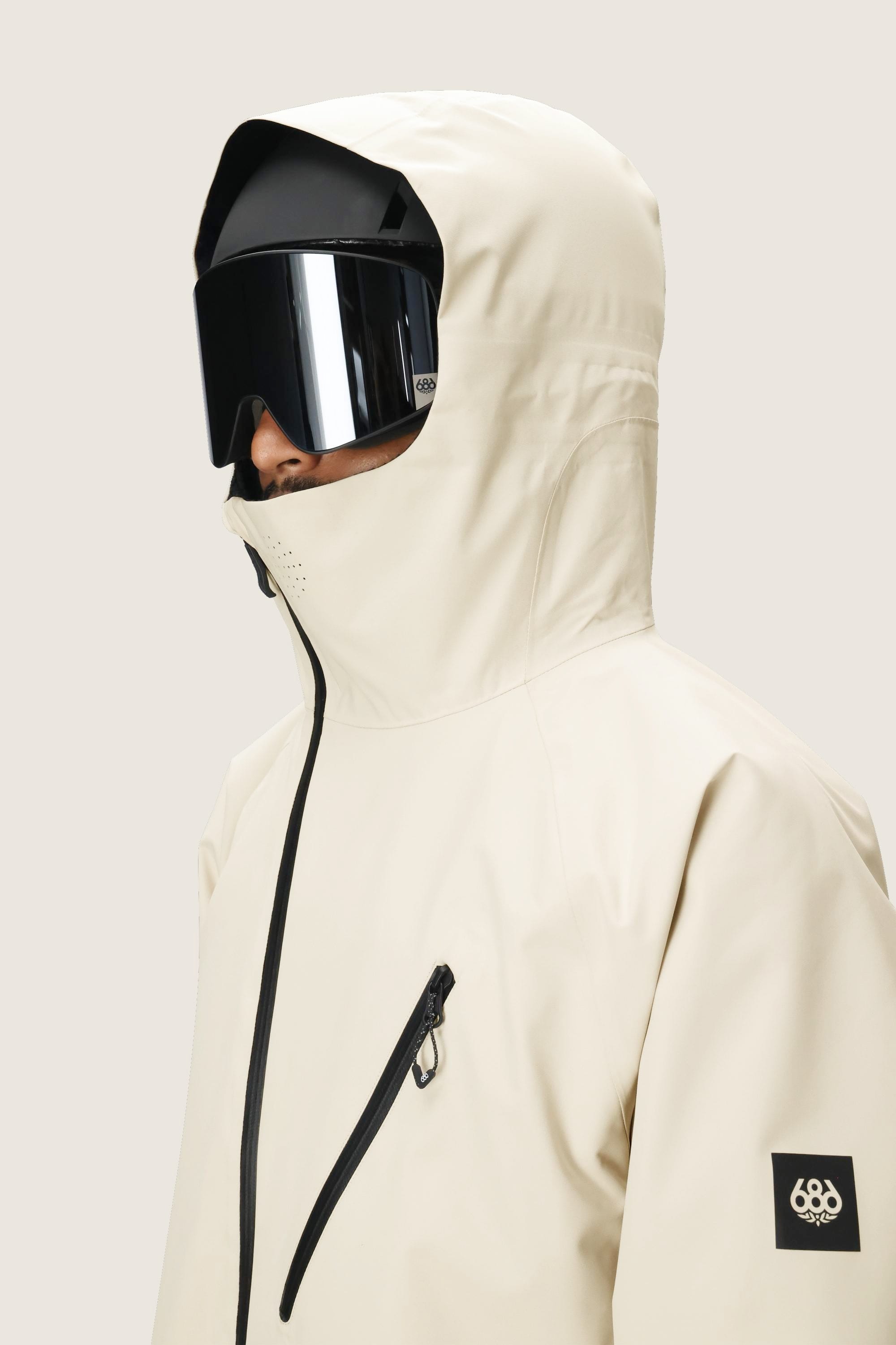 A person wearing the 686 Men's Hydra™ Thermagraph® Jacket in cream with a black front zipper, chest pocket, and 686 logo patch on the sleeve, plus large black ski goggles, is shown in a side view.