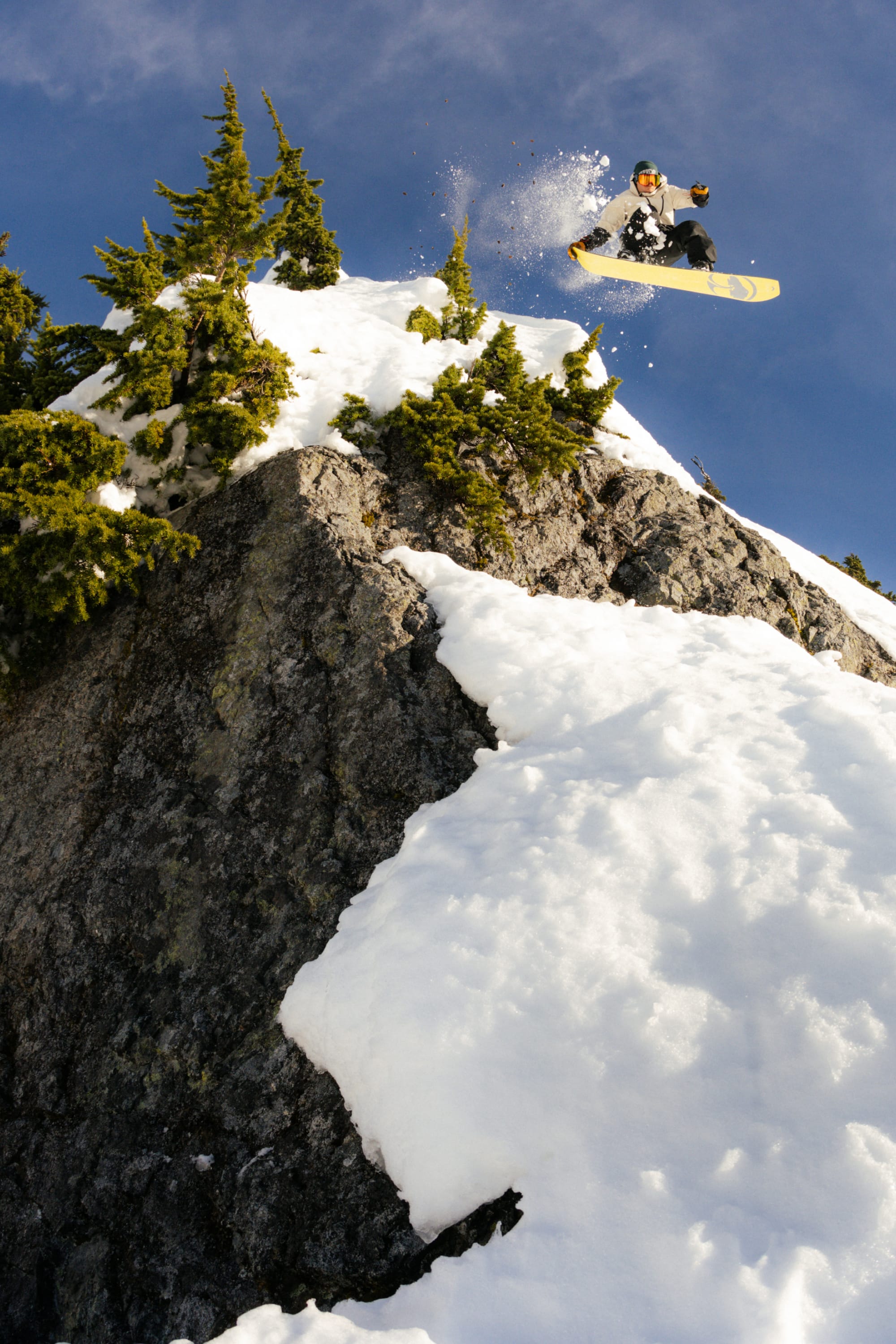A snowboarder in the 686 Men's Hydra™ Thermagraph® Jacket leaps off a snowy mountain cliff, evergreen trees and blue sky behind, while snow sprays in their wake.