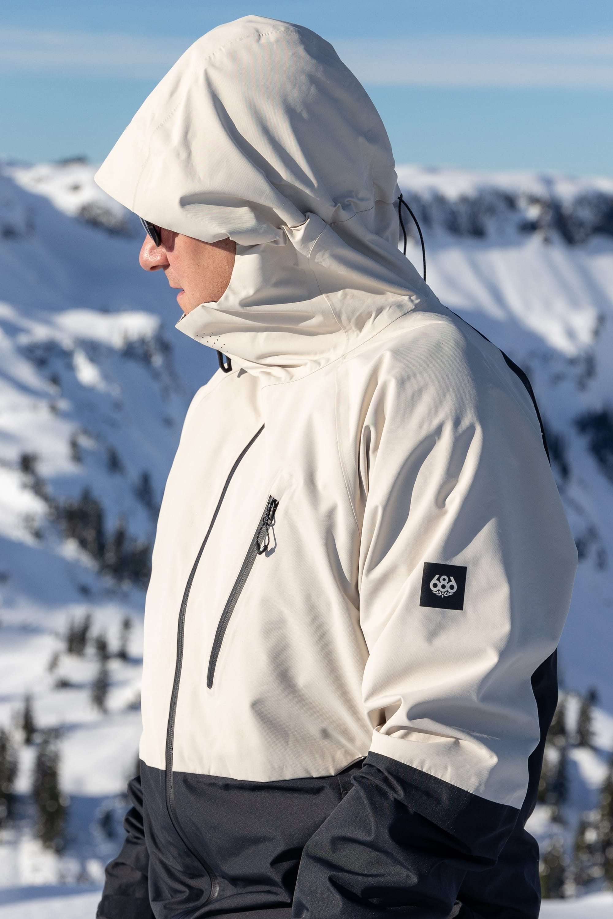 A person wearing the 686 Men's Hydra™ Thermagraph® Jacket, a hooded light-colored coat with body-mapped lining, stands sideways outdoors in snow-covered mountains under a clear sky.