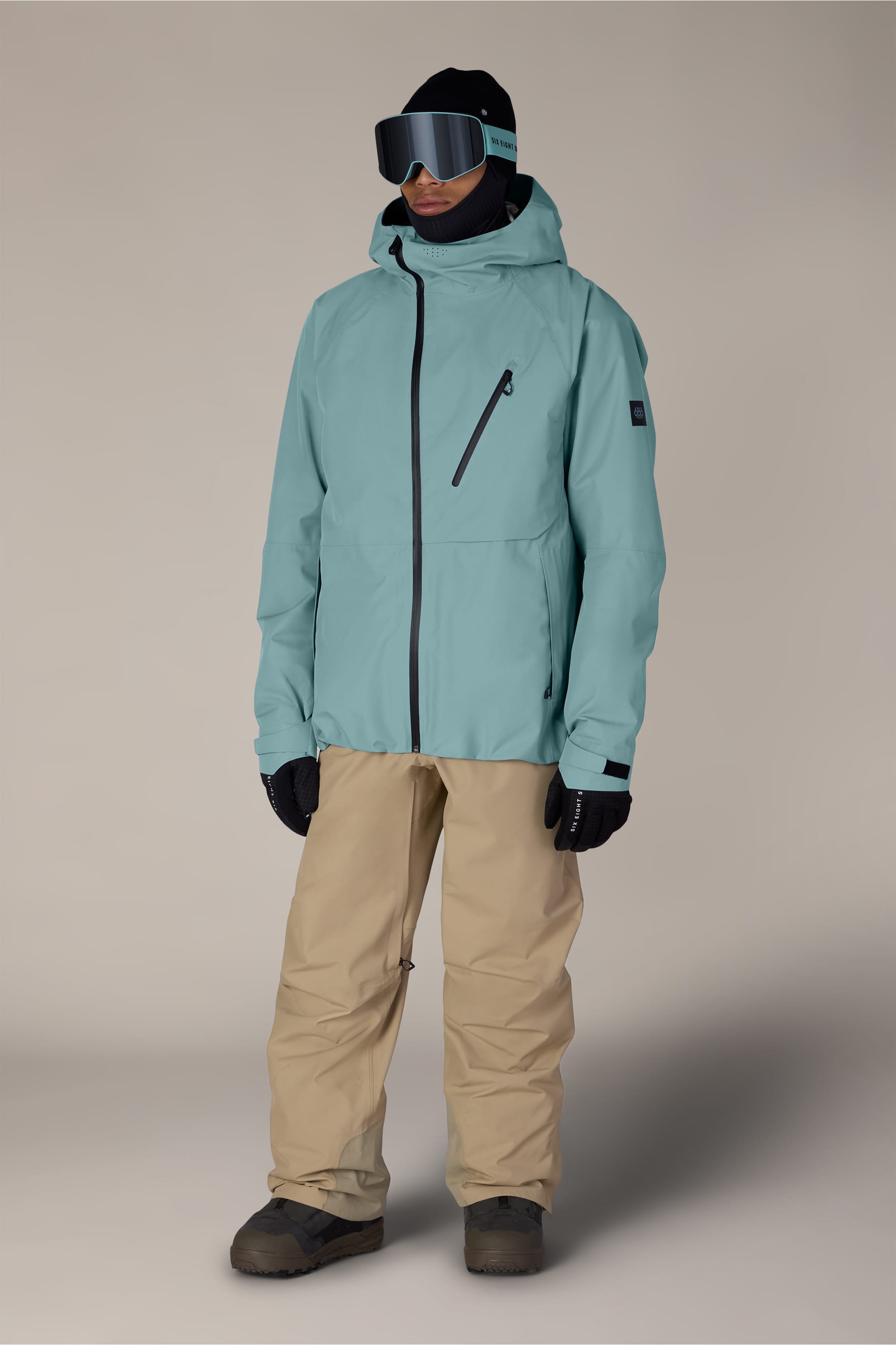 A person in ski gear stands facing forward, wearing the 686 Men’s Hydra™ Thermagraph® Jacket in light blue, paired with beige snow pants, black gloves, a black beanie, black ski goggles, and black boots against a plain light background.
