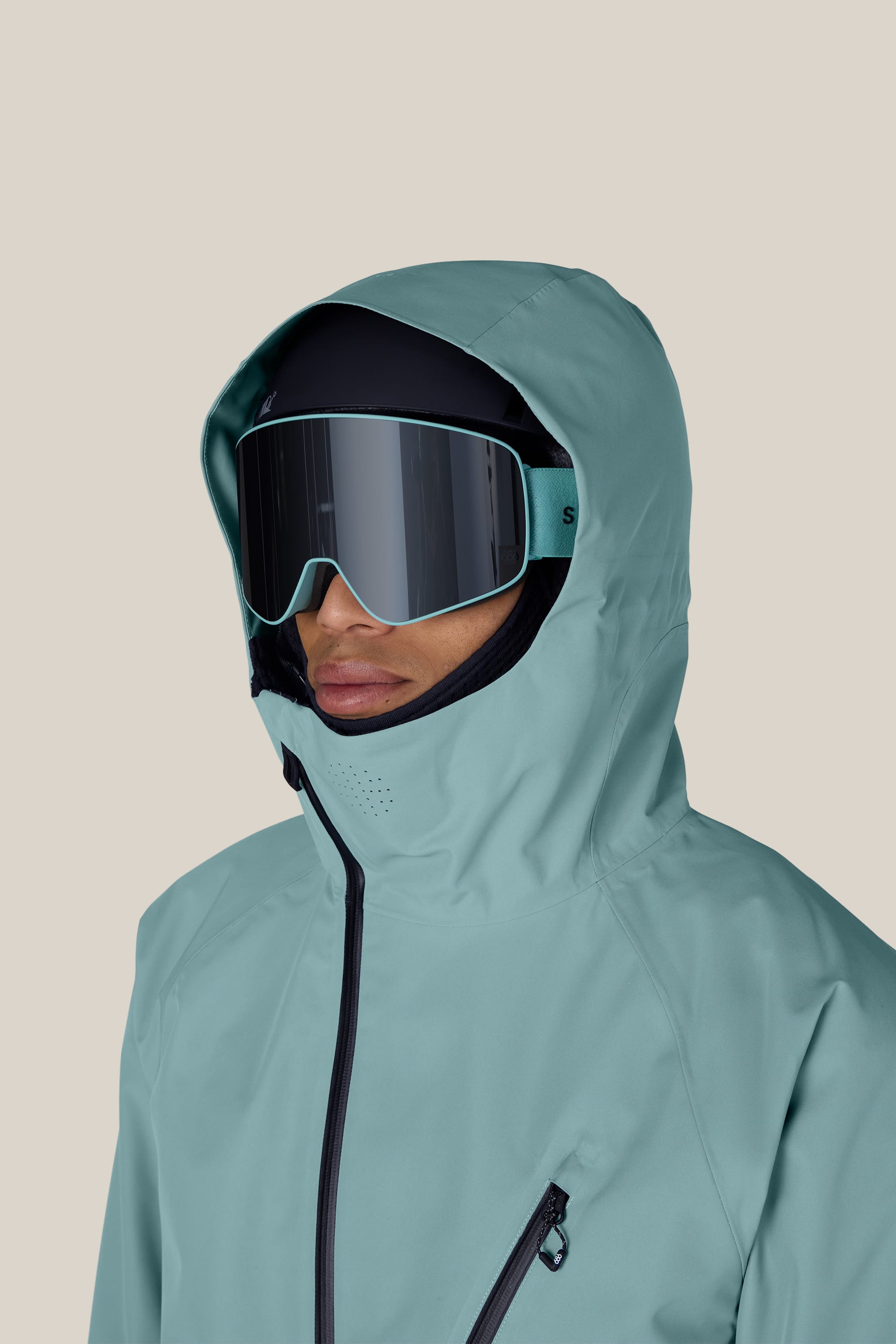 A person wearing the 686 Men’s Hydra™ Thermagraph® Jacket in teal, paired with black ski goggles and a helmet, faces slightly to the side against a plain beige background.