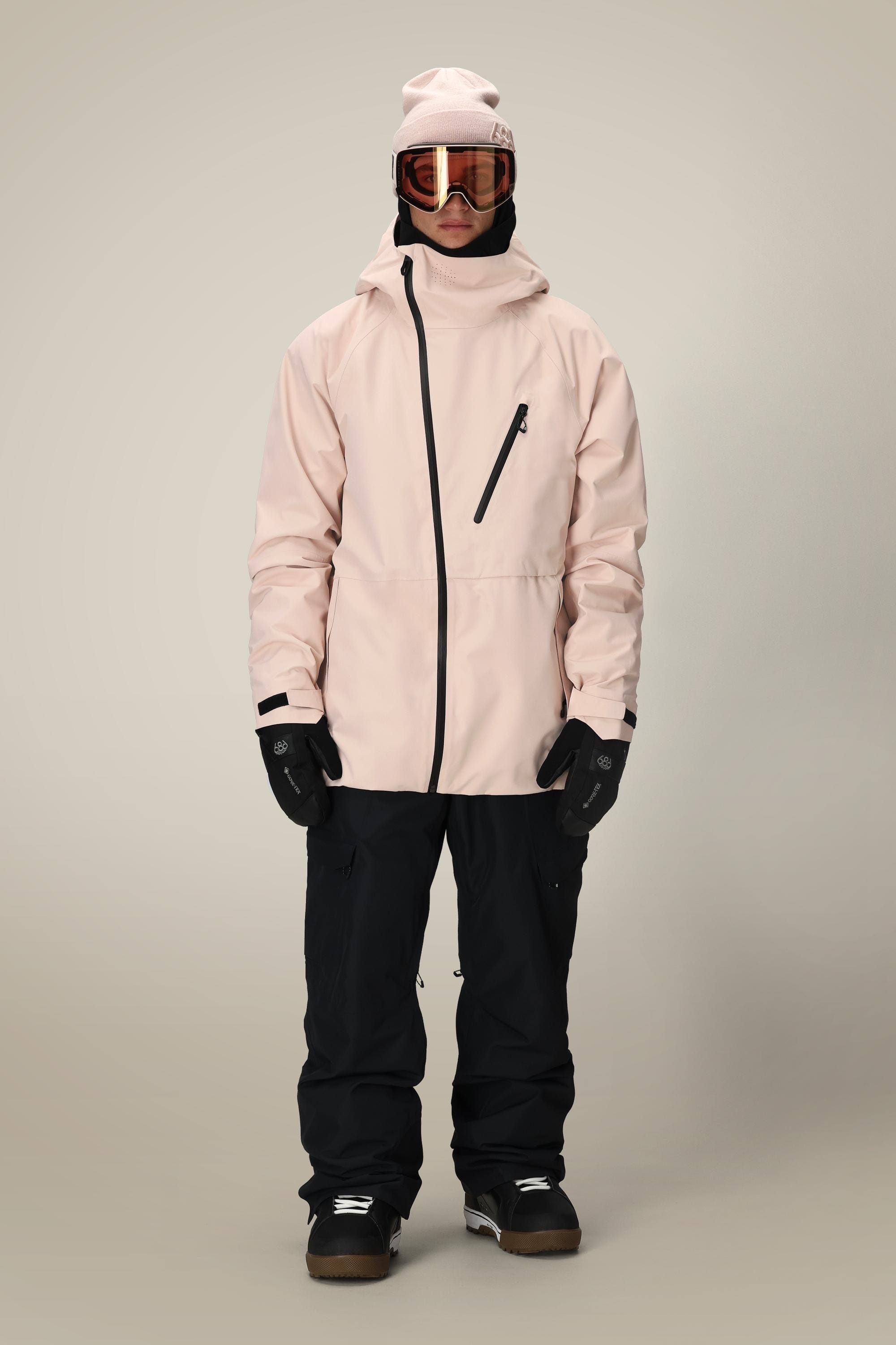 A person wearing the 686 Men's Hydra™ Thermagraph® Jacket in light pink, along with black snow pants, black gloves, goggles, and a beanie, stands facing forward against a plain light background.
