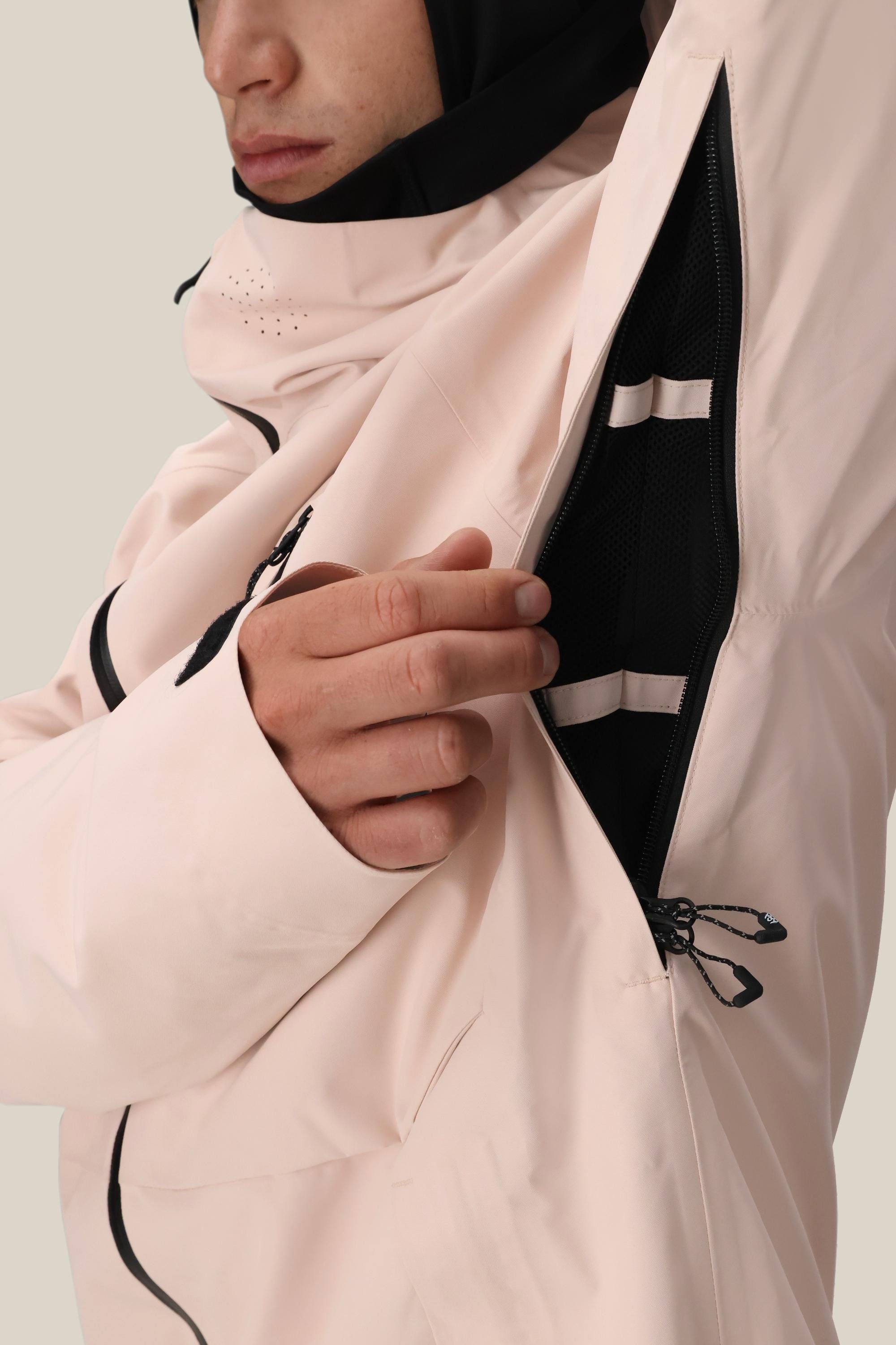 Wearing a pale pink 686 Men's Hydra™ Thermagraph® Jacket, a person unzips a black mesh-lined underarm vent to show interior ventilation, body-mapped lining, and drawstrings. Their hood is up and their face is partly visible.