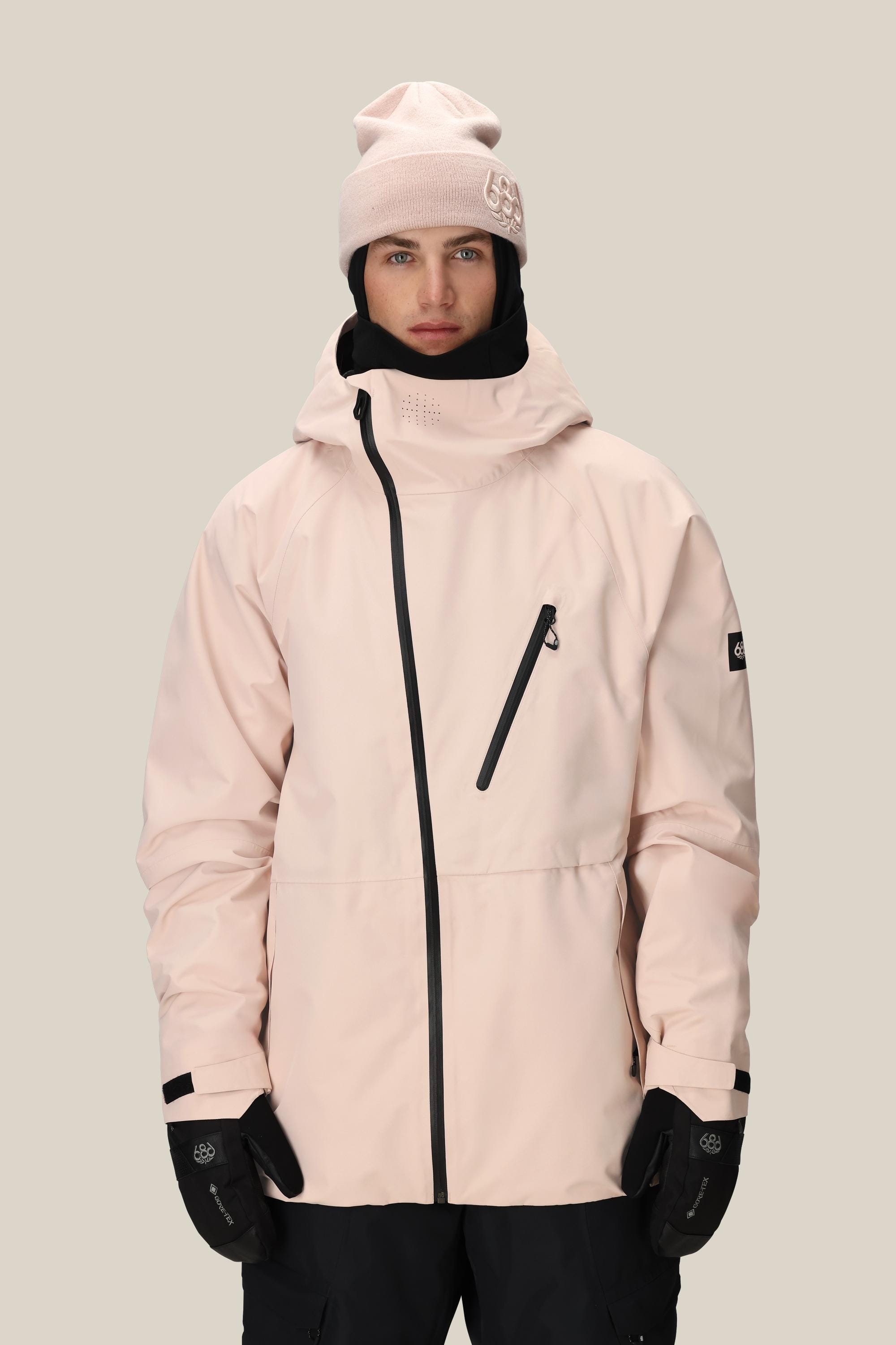 A person wears the 686 Men's Hydra™ Thermagraph® Jacket in light pink with black zippers, a matching pink beanie, black gloves, and black pants while standing against a plain beige background.