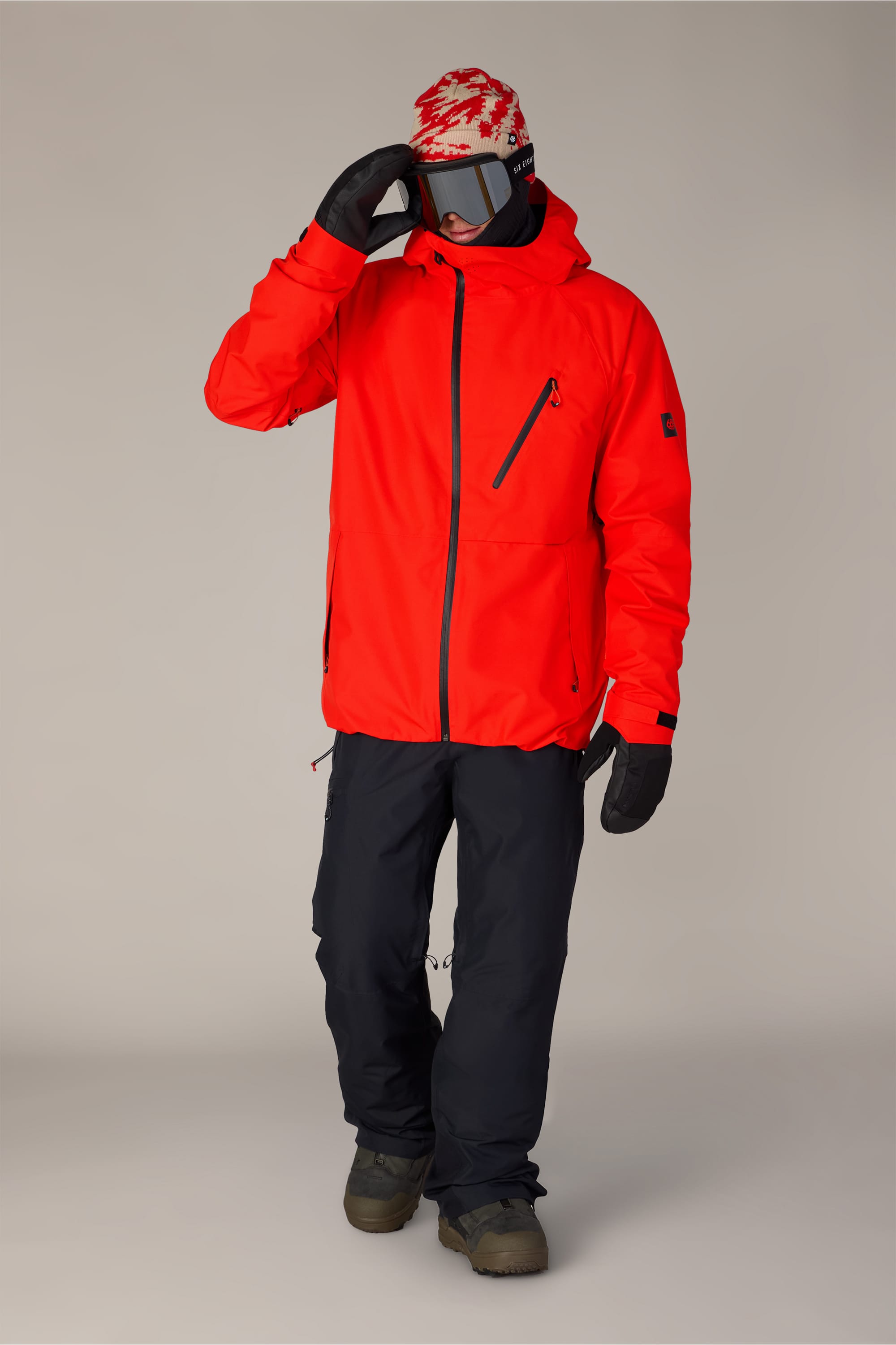 A person wearing the 686 Men’s Hydra™ Thermagraph® Jacket in bright red, paired with black pants, black gloves, winter boots, a red-and-white beanie, and ski goggles stands against a plain background, lifting one hand toward their goggles.