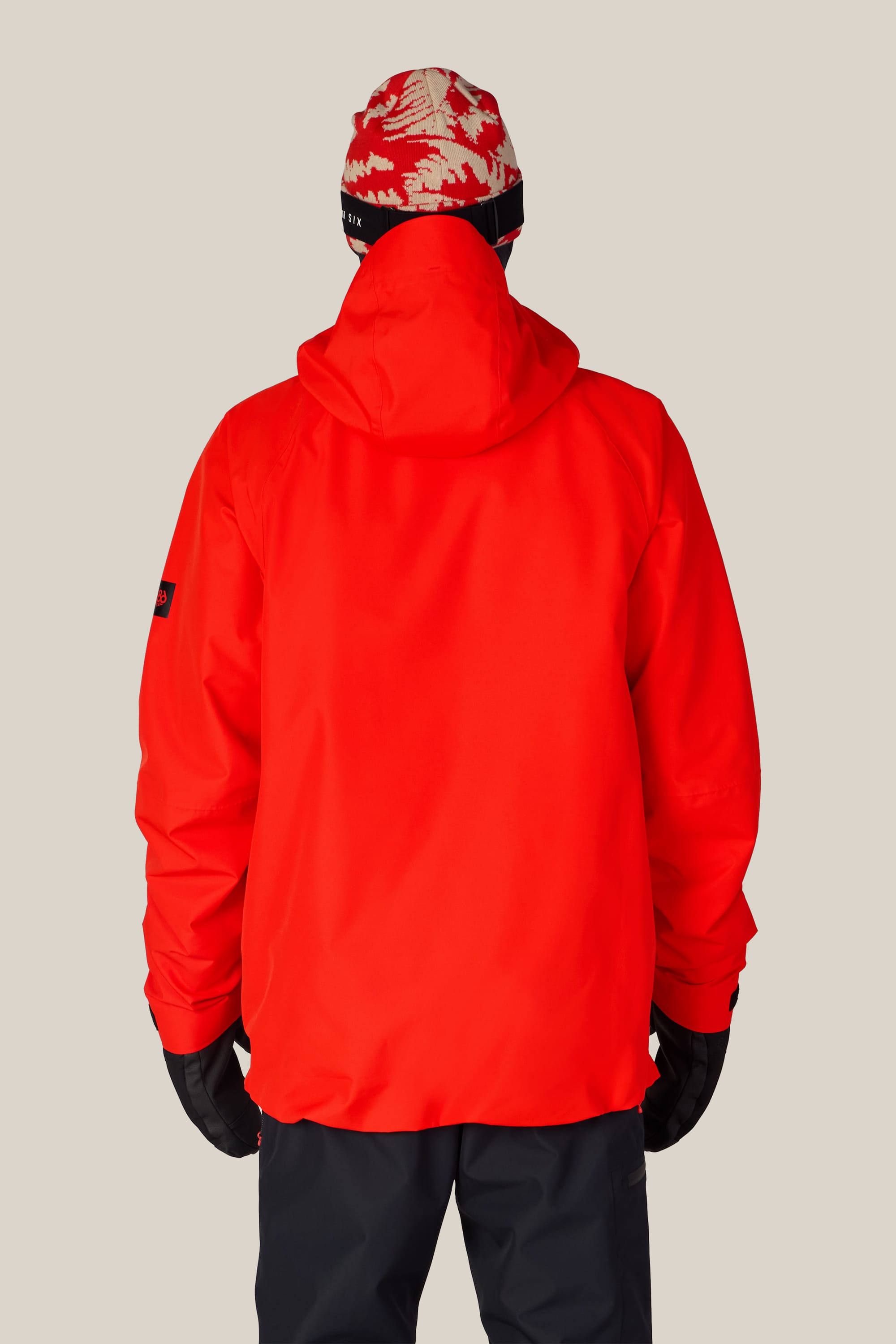 A person seen from the back, wearing the 686 Men’s Hydra™ Thermagraph® Jacket in bright red by 686, along with a patterned red and white beanie, black gloves, and dark pants, standing against a plain light background.