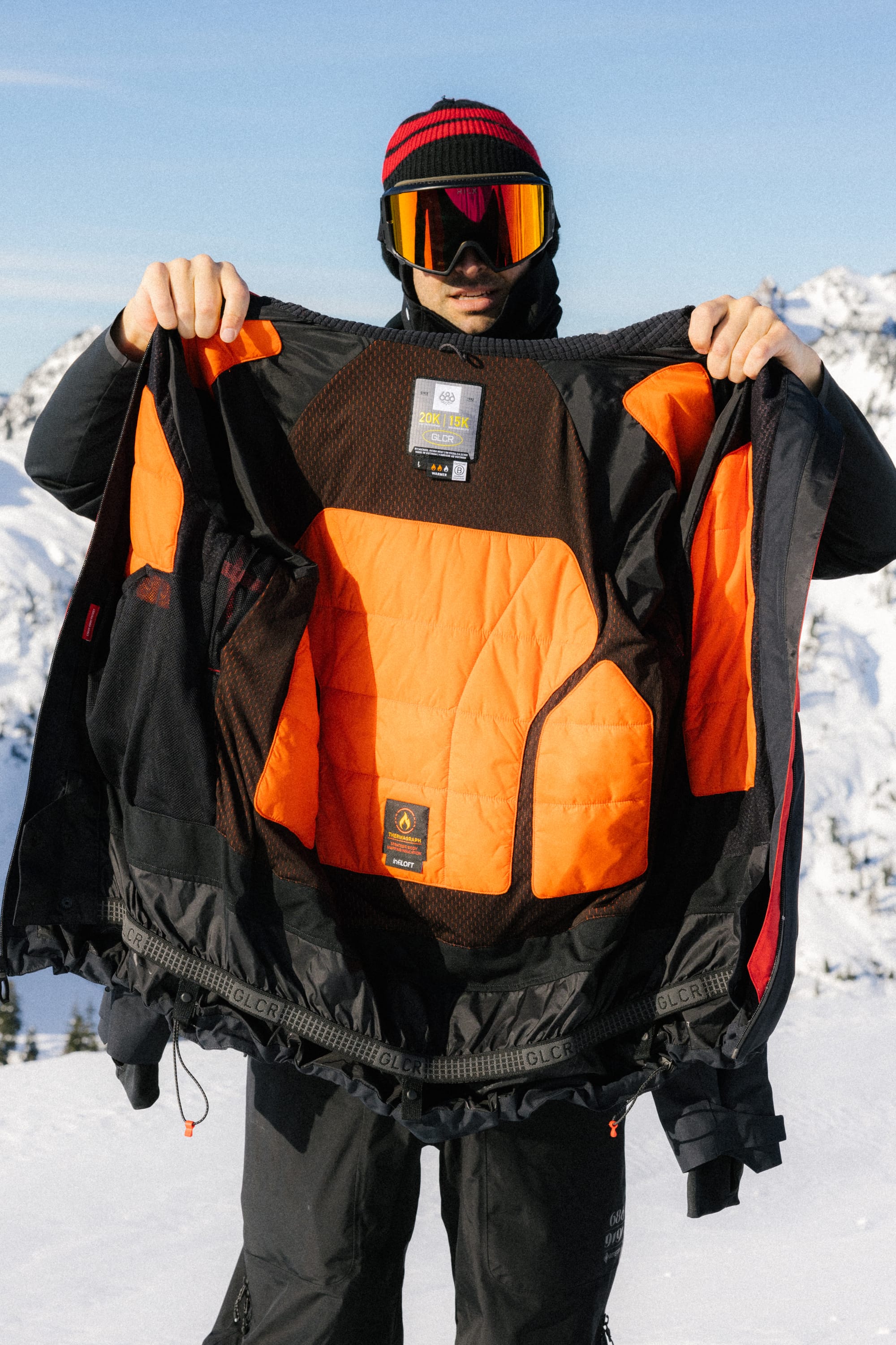 A person in ski gear stands on a snowy mountain, holding open the 686 Men's Hydra™ Thermagraph® Jacket to reveal its body-mapped lining and labels with snow-covered peaks in the background.