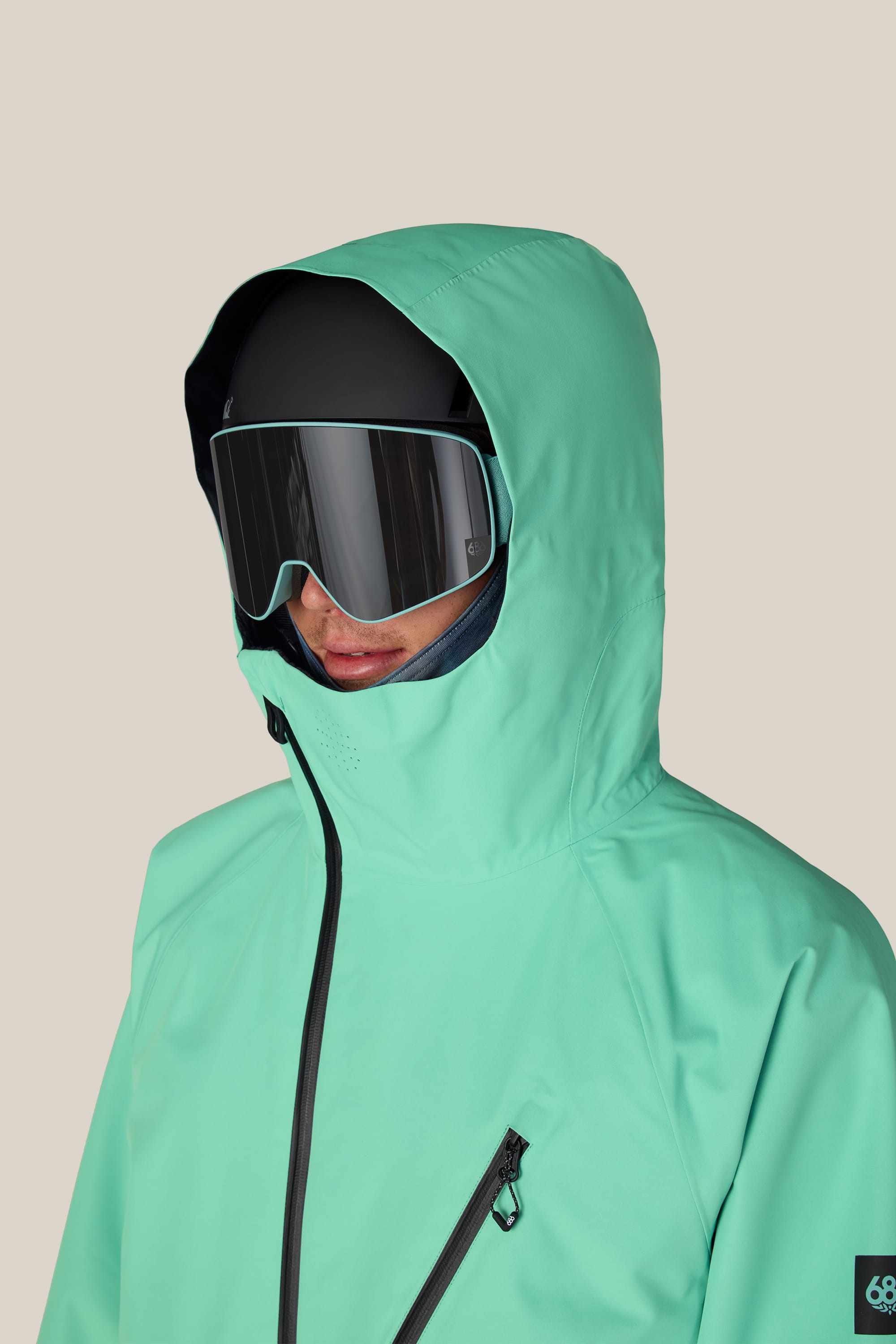 A person wearing the 686 Men’s Hydra™ Thermagraph® Jacket in mint green, paired with a black ski helmet and reflective ski goggles, stands against a plain beige background.