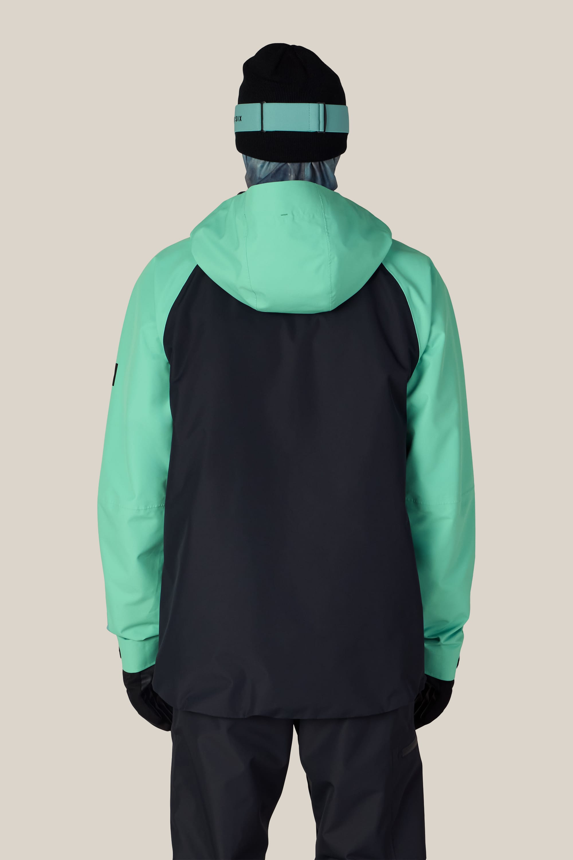 A person shown from the back against a plain beige background is wearing the 686 Men’s Hydra™ Thermagraph® Jacket by 686 in black and teal, paired with black pants, gloves, and a black beanie featuring a teal stripe.