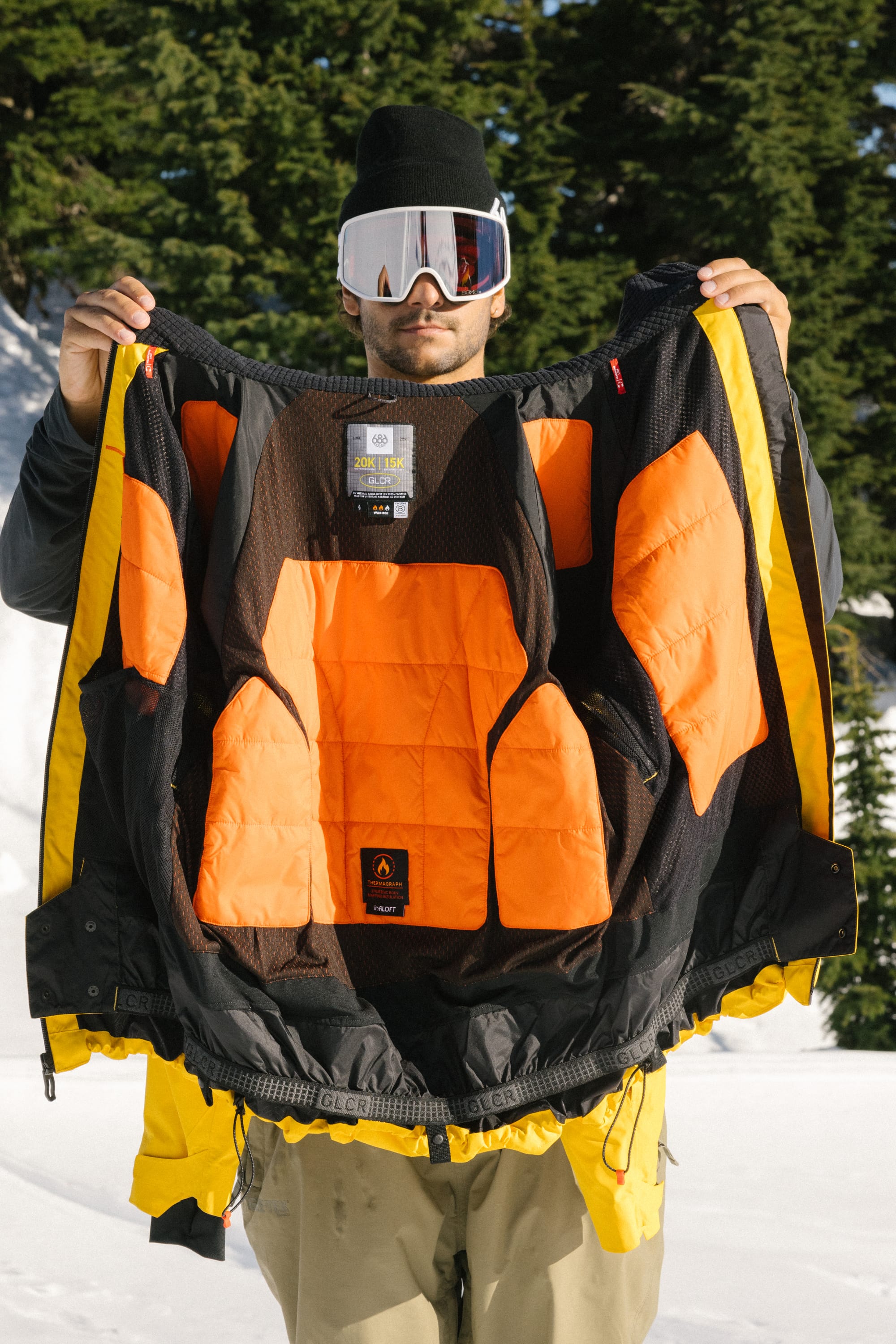 A person in 686 ski goggles and a black beanie holds open the 686 Men's Hydra™ Thermagraph® Jacket, revealing its orange, body-mapped lining against a snowy backdrop with evergreen trees.