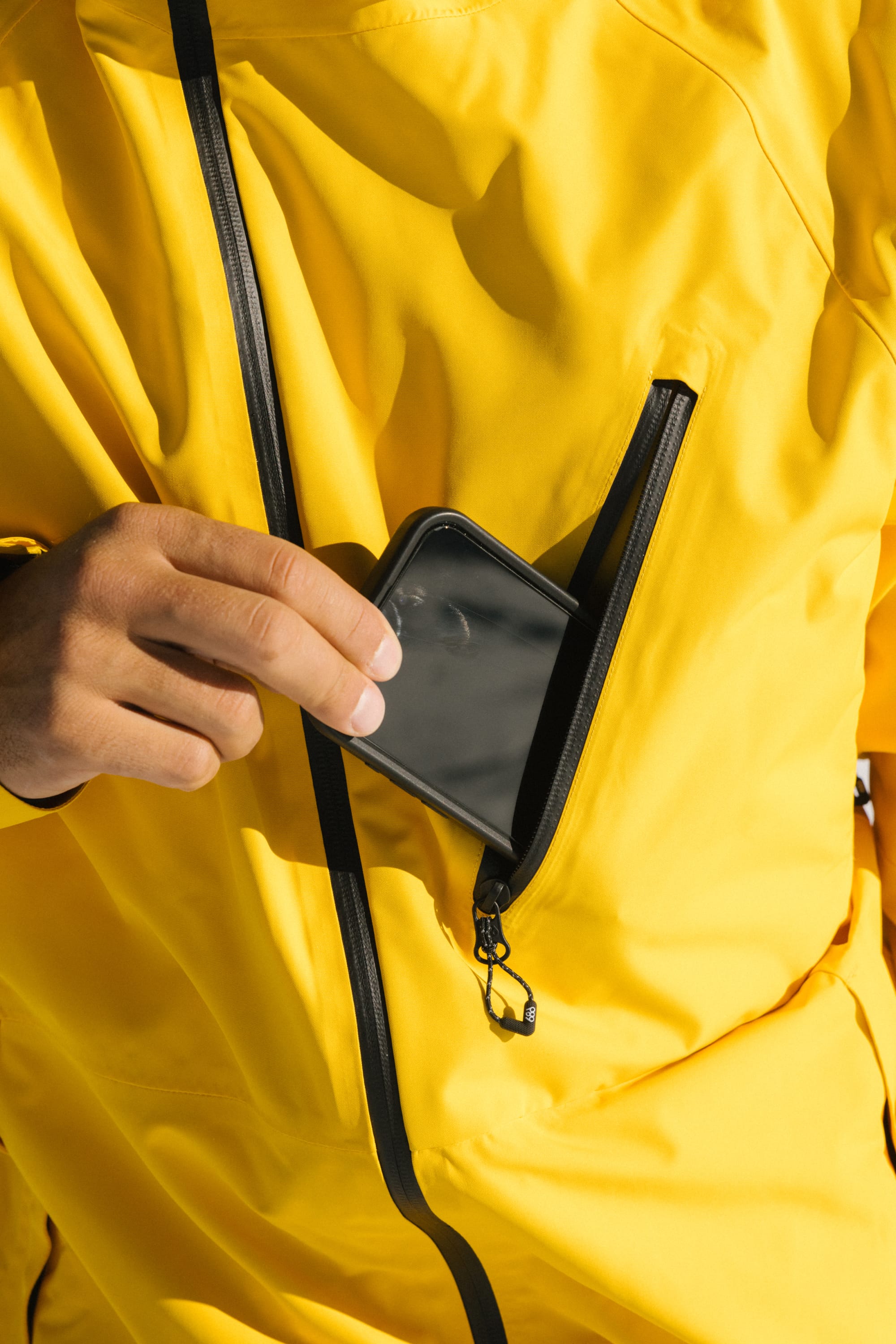 Wearing the 686 Men's Hydra™ Thermagraph® Jacket, a person slips a black smartphone into the zippered chest pocket of the bright yellow, body-mapped lined jacket by 686.
