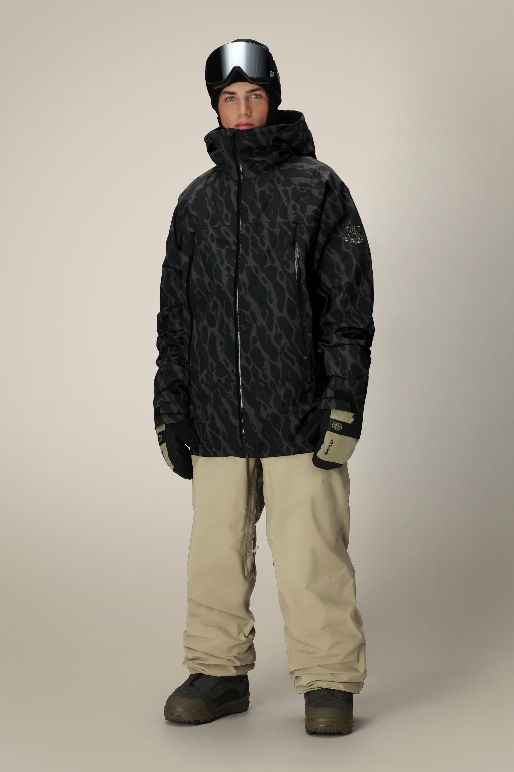A person wearing a 686 Men's SMARTY® 3-In-1 Rodeo™ 3L Jacket, beige snow pants, black gloves, gray snow boots, and black goggles stands against a plain light background.
