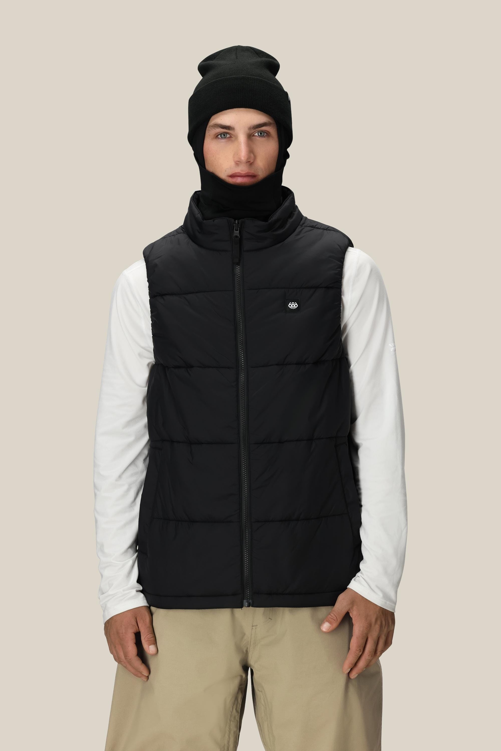 A person wearing a black beanie, the 686 Men's SMARTY® 3-In-1 Rodeo™ 3L Jacket over a white long-sleeve shirt, and beige pants stands against a plain light beige background.