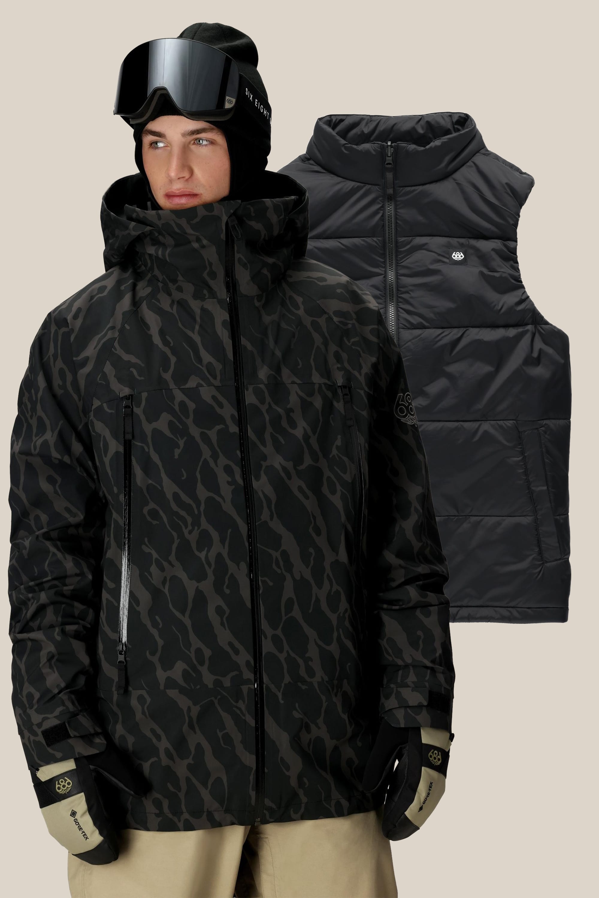 A person wears the 686 Men's SMARTY® 3-In-1 Rodeo™ 3L Jacket in black and gray camo with black gloves, tan pants, a black beanie, and ski goggles. Behind them is a matching black 686 puffer vest on a beige background.
