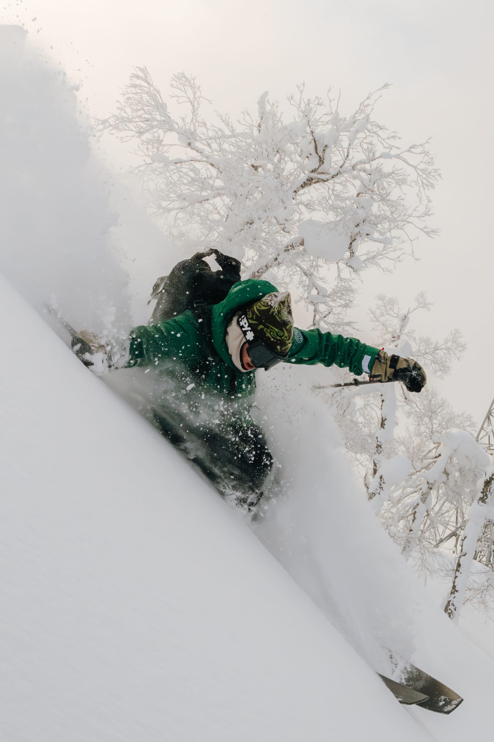 A snowboarder wearing the 686 Men's SMARTY® 3-In-1 Rodeo™ 3L Jacket and helmet carves through deep powder on a snowy slope, with snow-covered trees in the background.