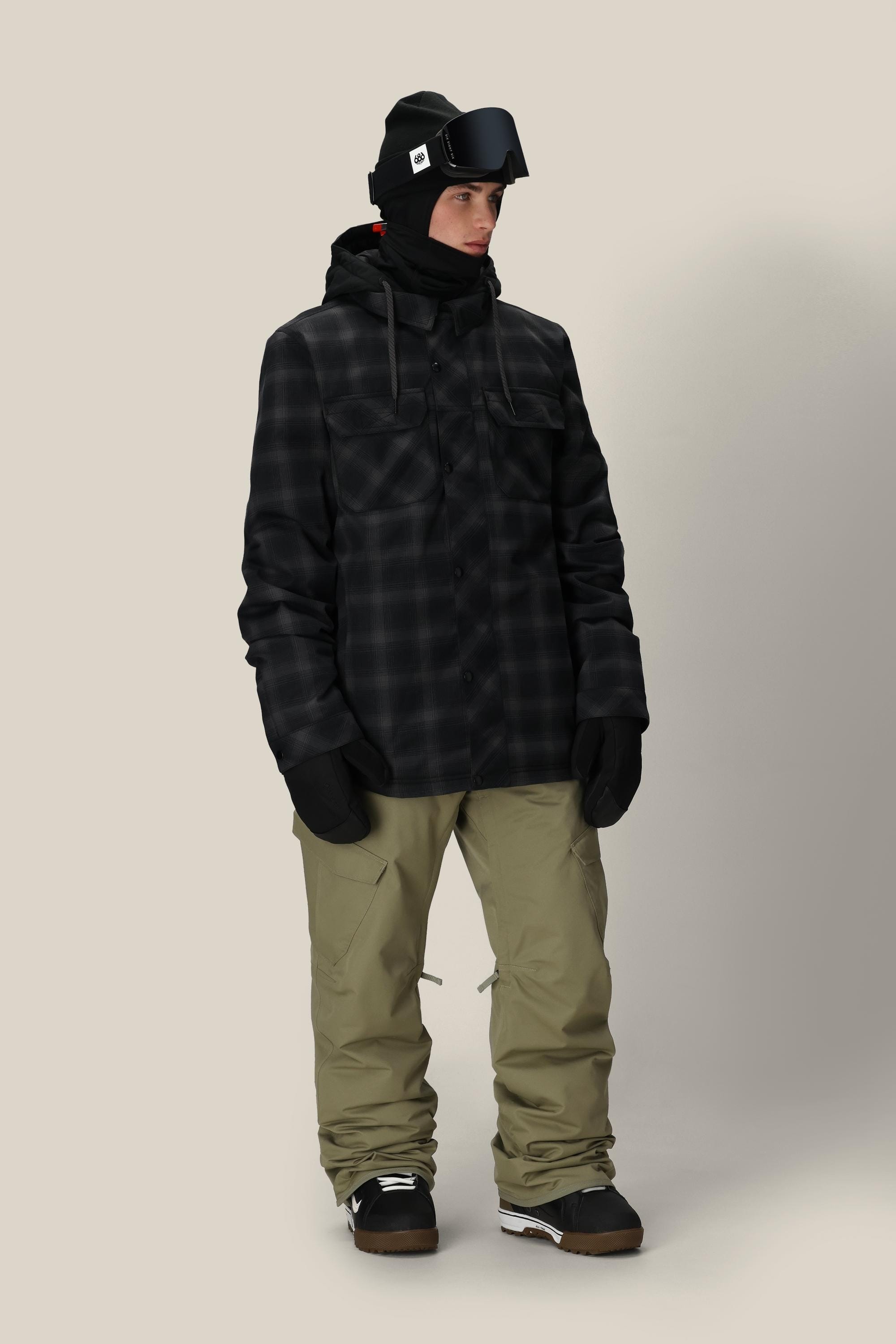 A person wearing a 686 Men's Woodland™ Insulated Jacket in black plaid, khaki snow pants, black gloves, winter boots, a black balaclava, and black ski goggles stands against a plain light background.