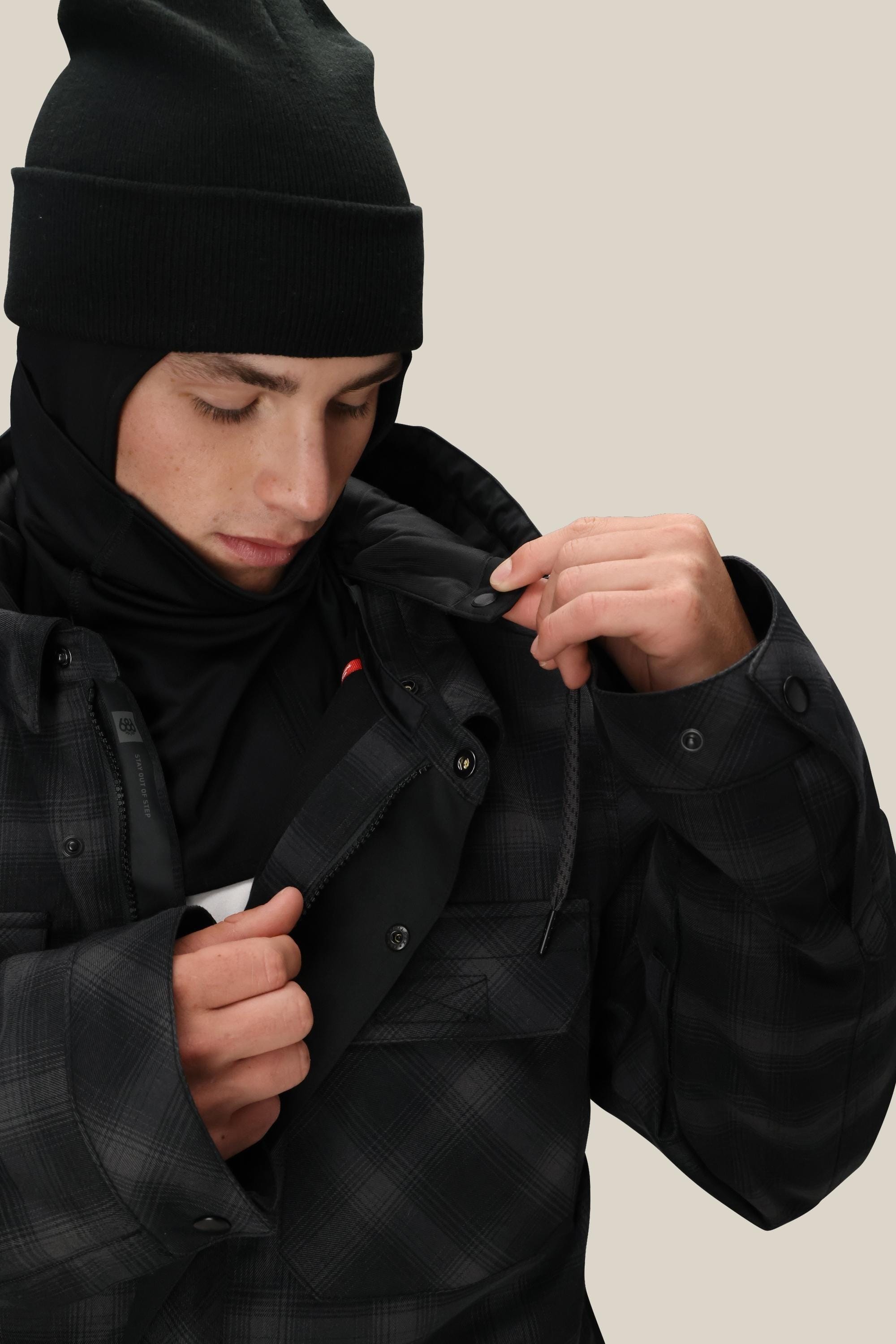 A man wearing a black beanie and 686 Men's Woodland™ Insulated Jacket by 686 looks down, unzipping the polyester waterproof jacket to reveal a black inner layer against a neutral background.